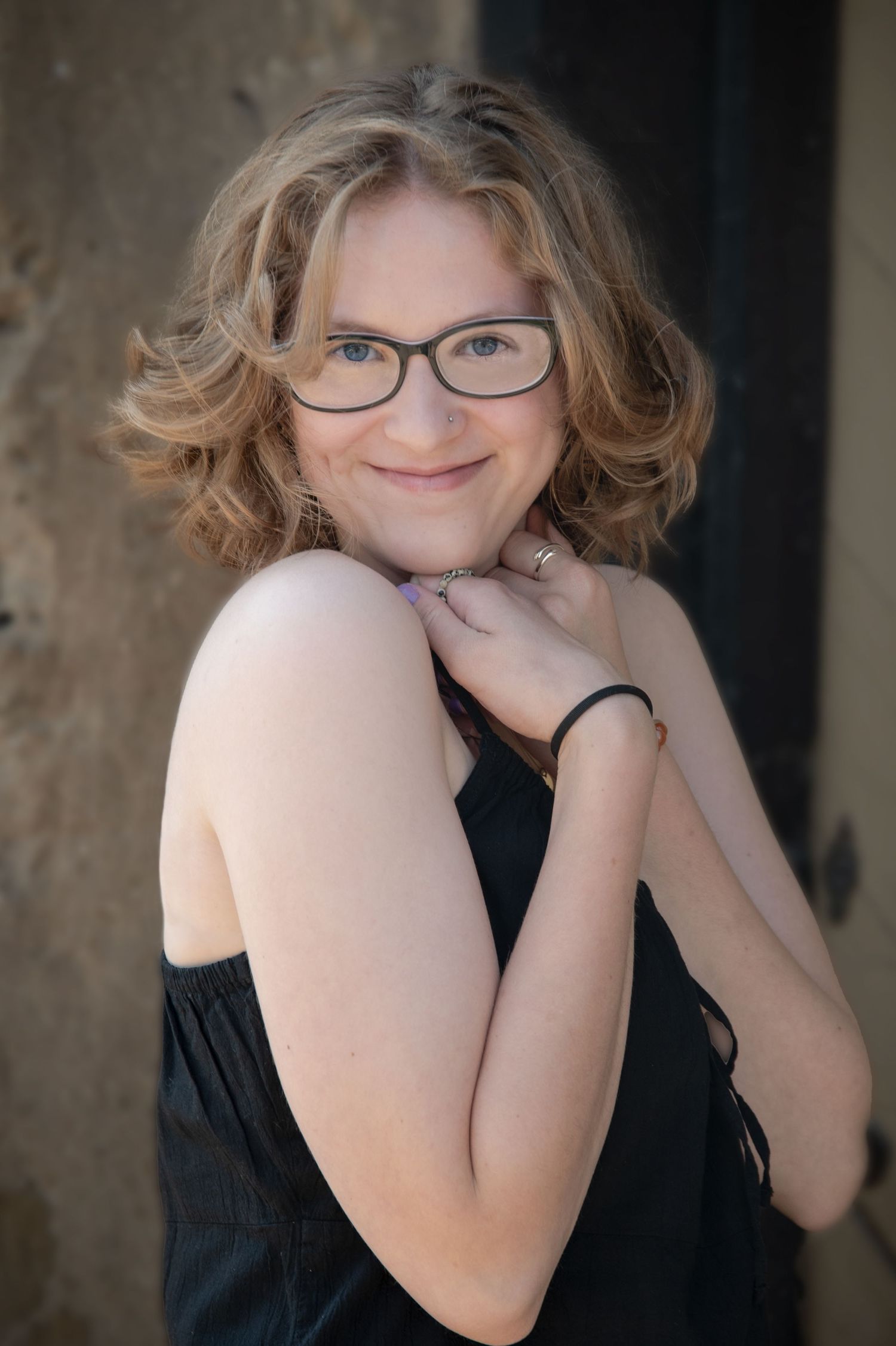 Young woman with curly blonde hair and glasses smiling over shoulder in black dress portrait.