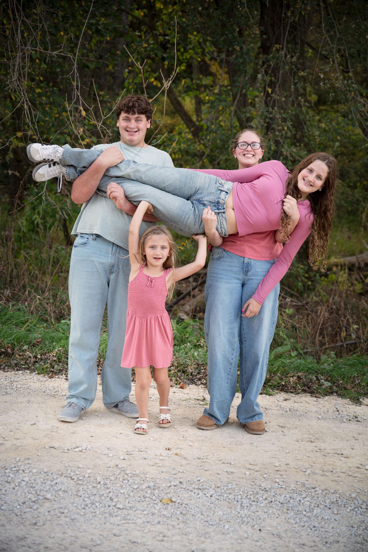 Four siblings laugh as two carry a girl horizontally on a gravel path outdoors.