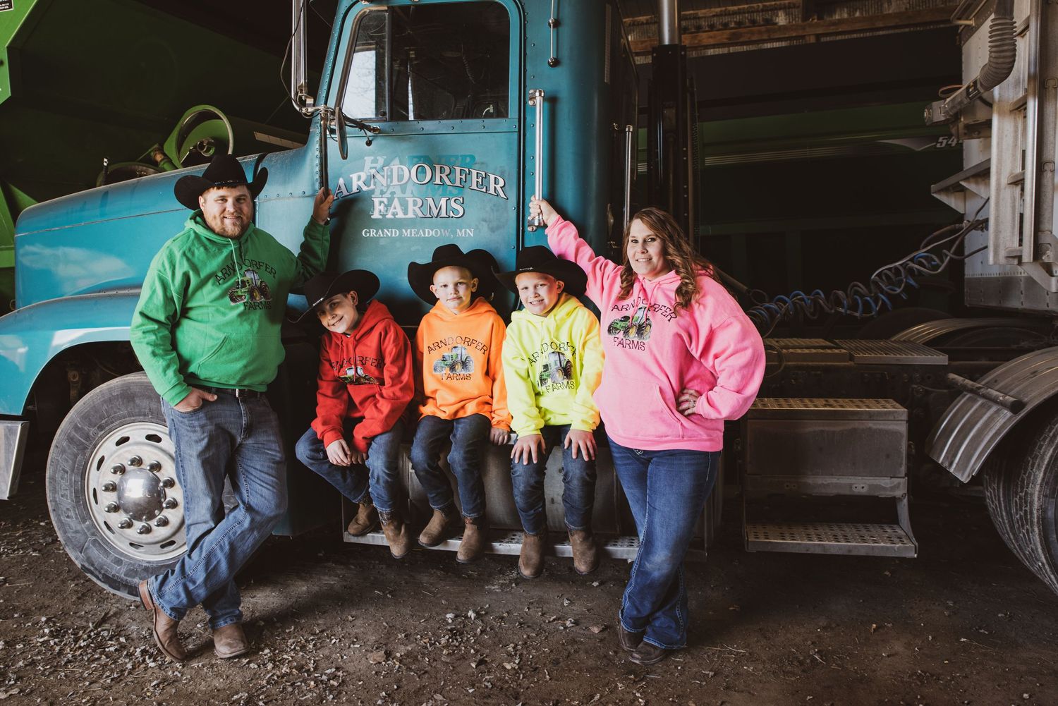Farm family of five poses in colorful hoodies beside a blue Porter Farms truck in a rustic barn setting.