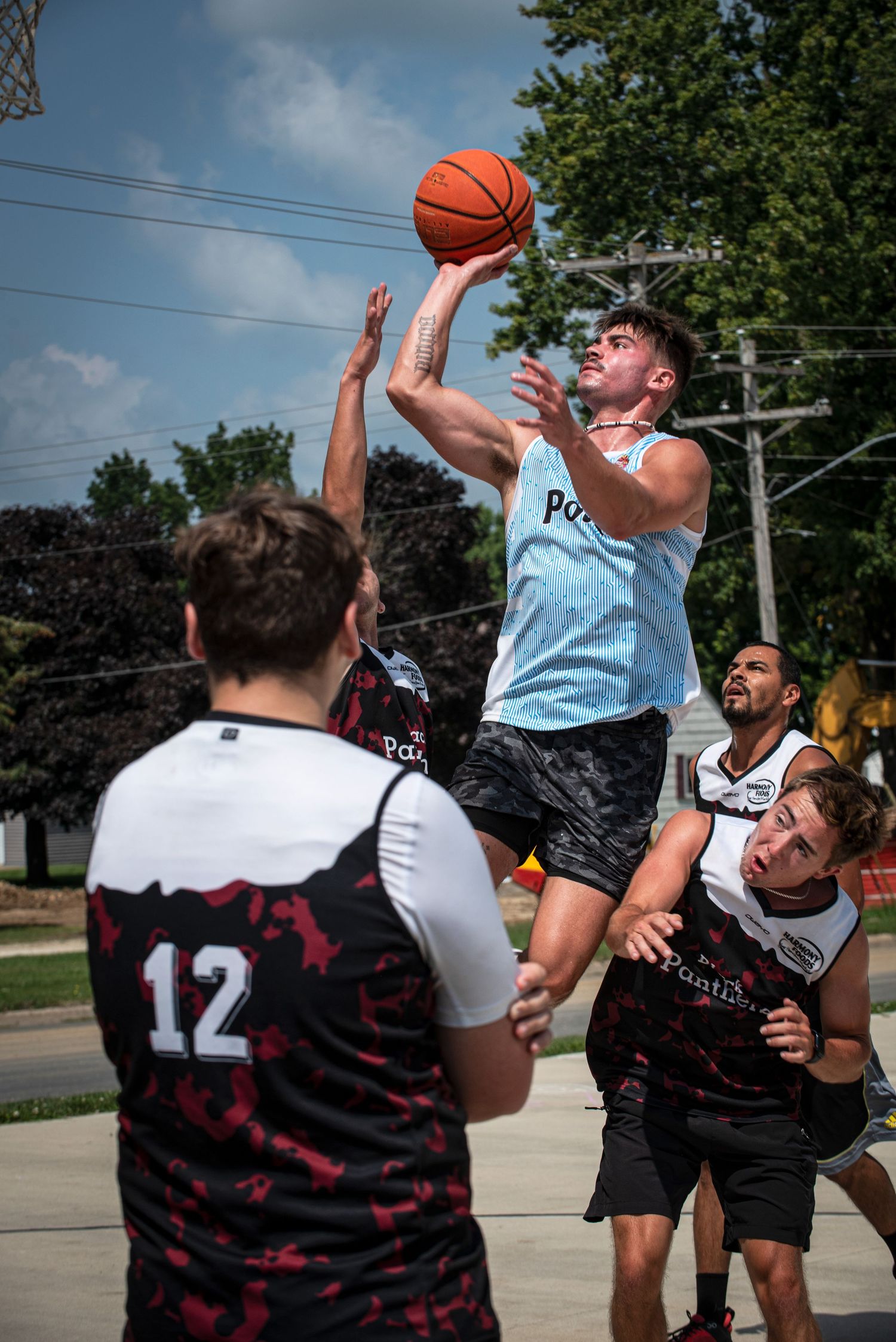 Athletic player in blue jersey leaps for layup surrounded by defenders in outdoor Rucker League basketball game.