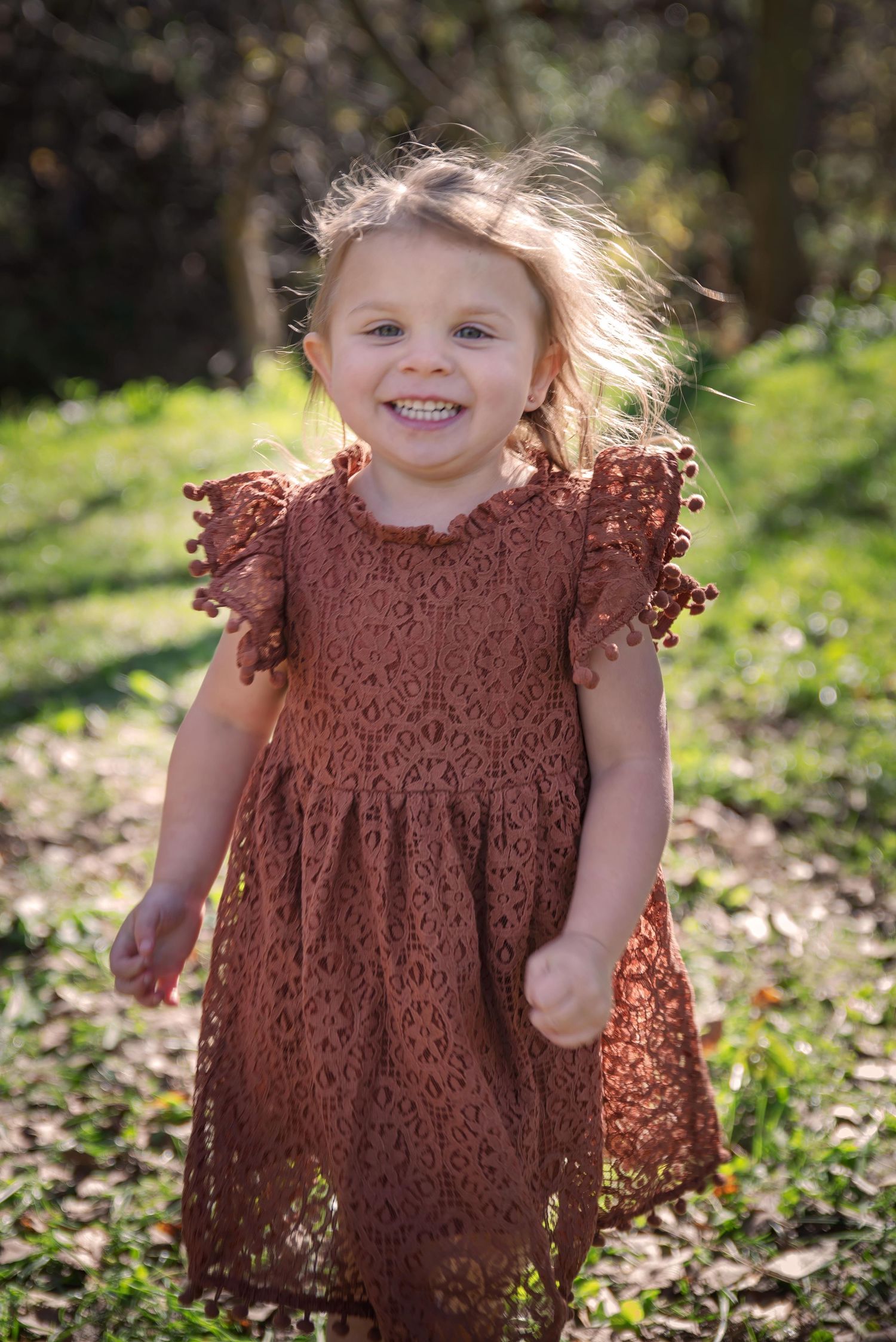 Happy toddler girl in rust-colored lace dress laughing outdoors in sunny park.