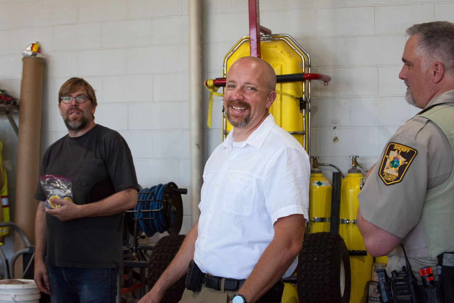Three men stand in a fire station garage, one wearing a sheriff uniform, smiling and conversing. 4th of July Celebration in Harmony MN.