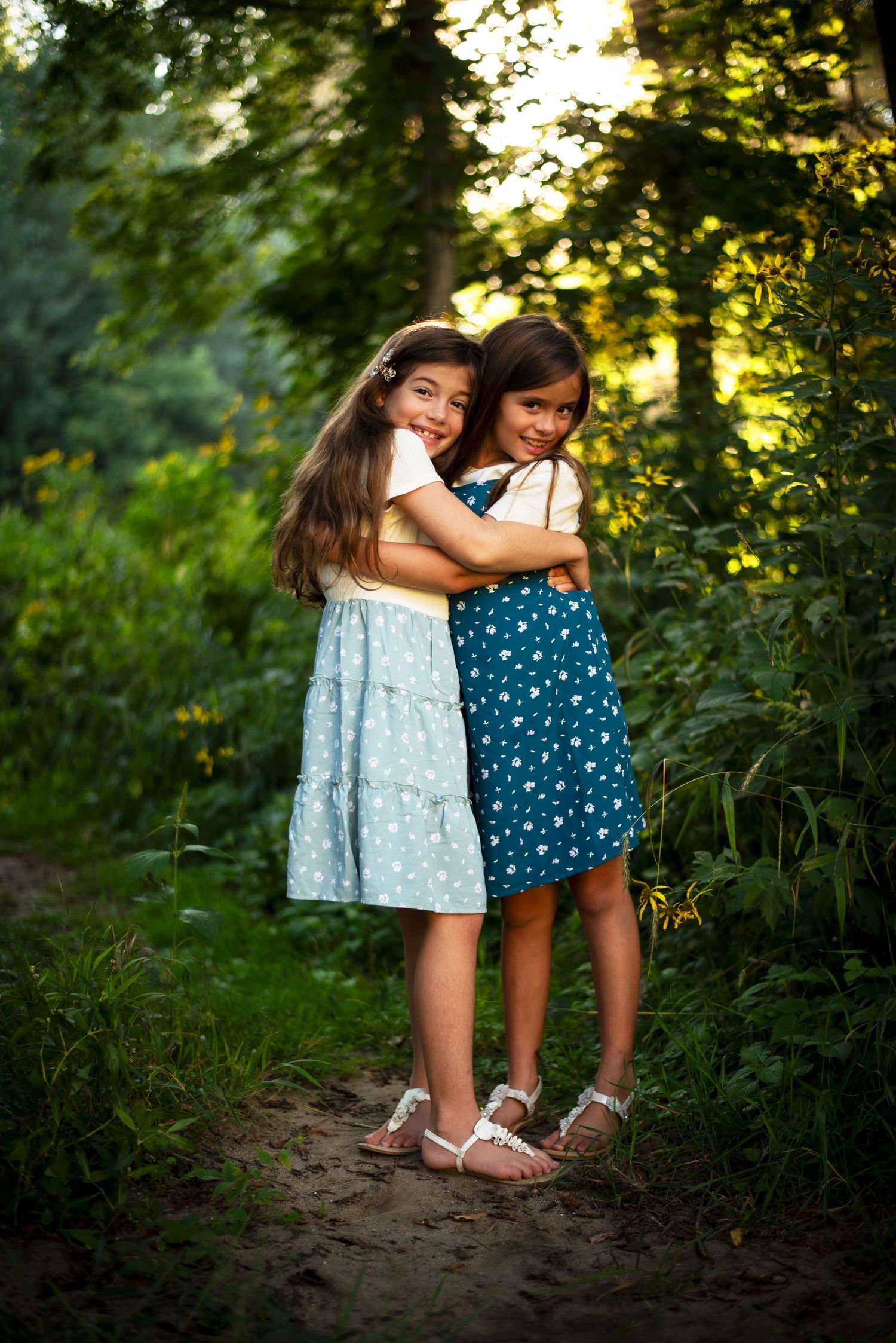 Two young girls hugging each other on a sunlit forest path surrounded by greenery.