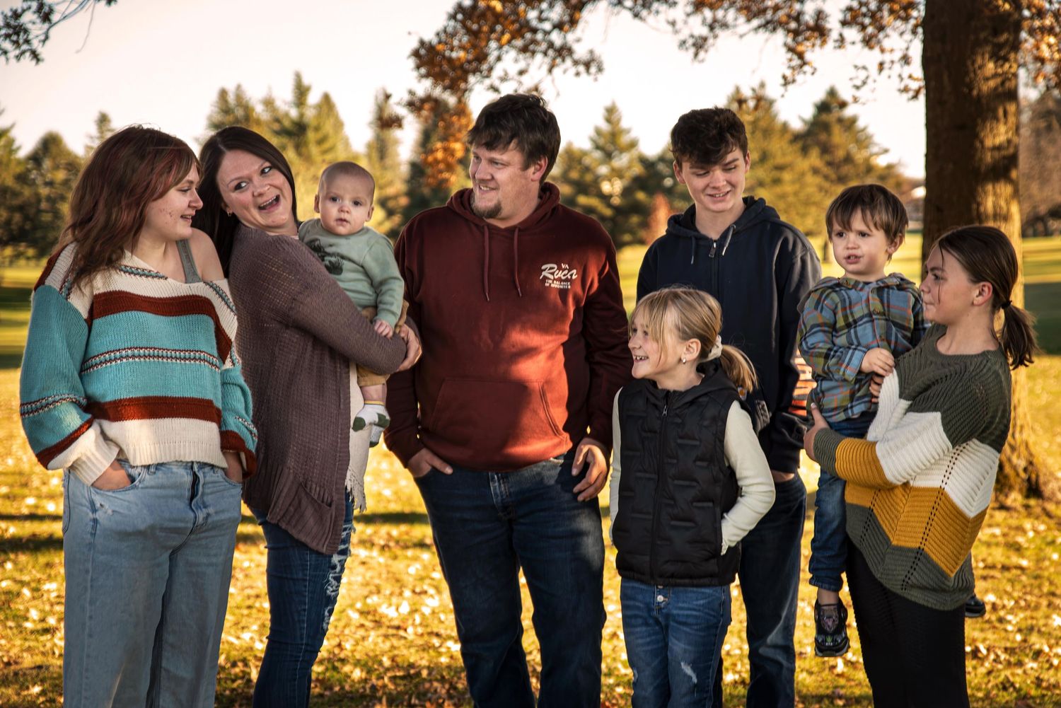 A large happy family laughs together outdoors among golden autumn fallen leaves.