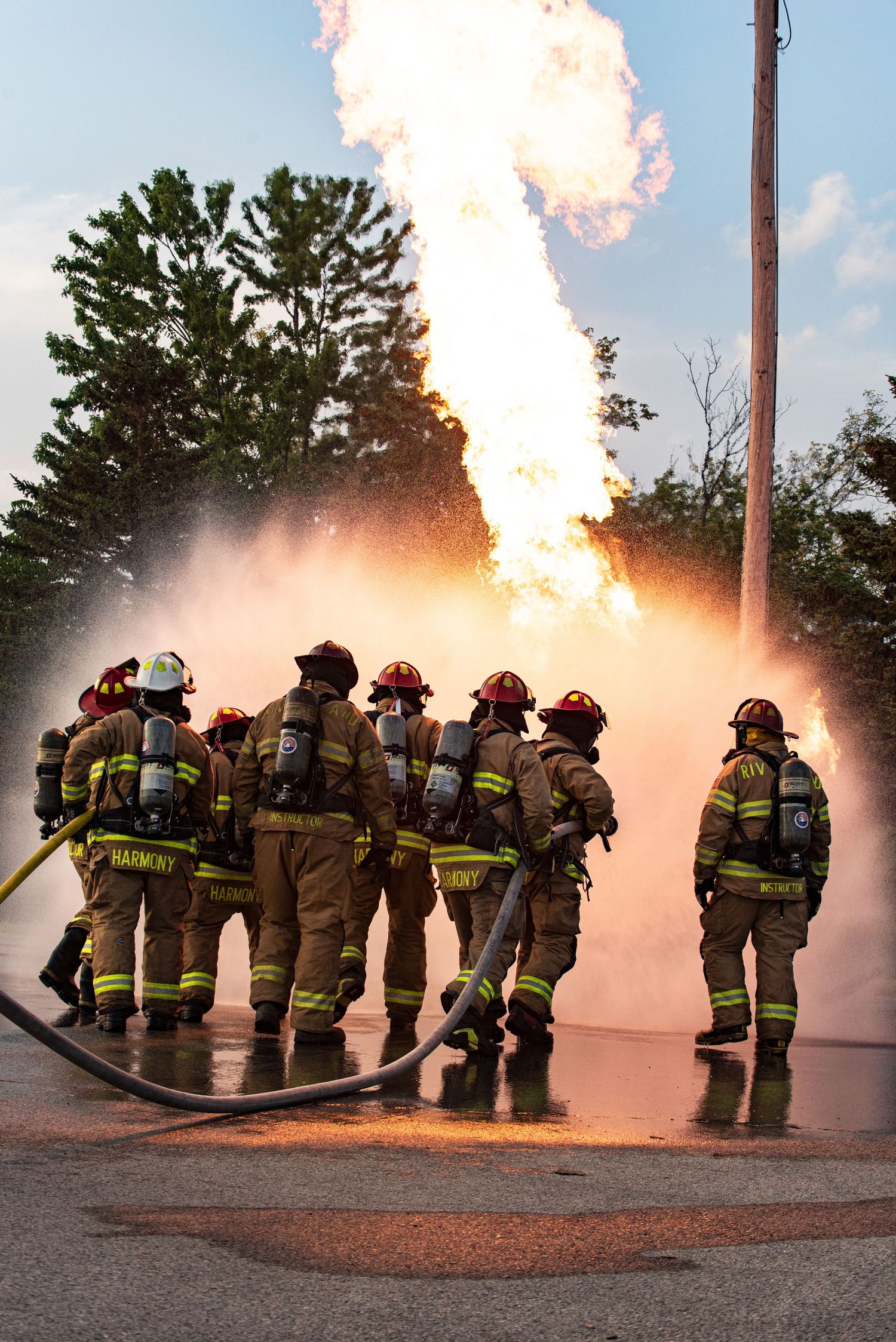 Firefighters in protective gear working together with hoses during a live fire training exercise.