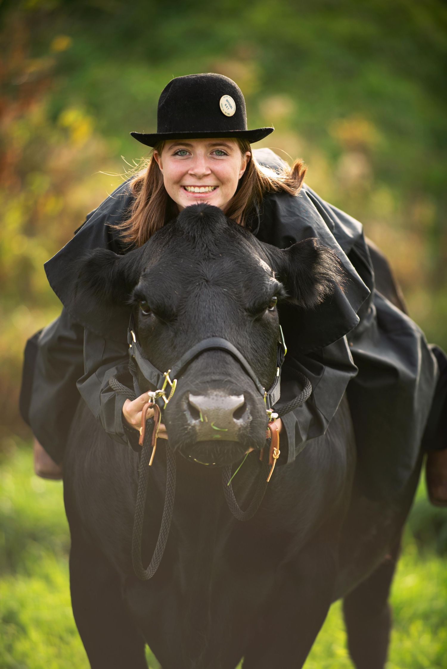 Smiling woman in black hat and cape leaning over a black bull in a green field.