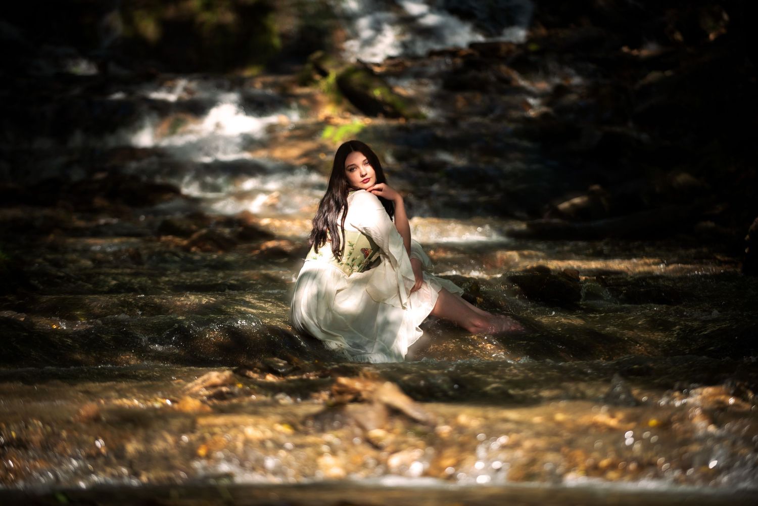 Young woman in white dress sitting gracefully in a flowing rocky stream surrounded by nature.