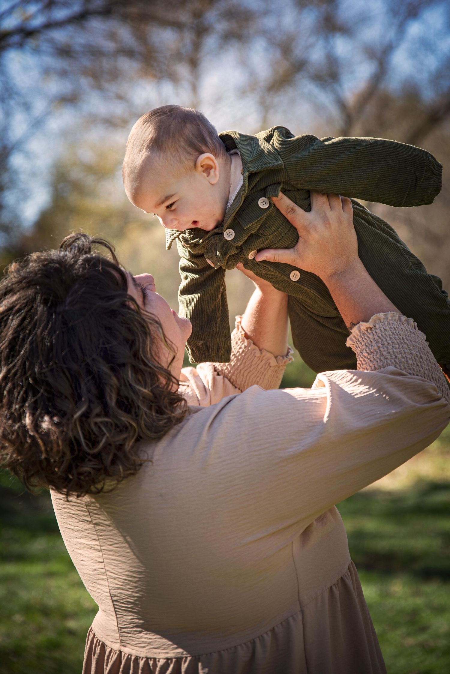 A mother joyfully lifts her baby in the air outdoors in a sunny park setting.
