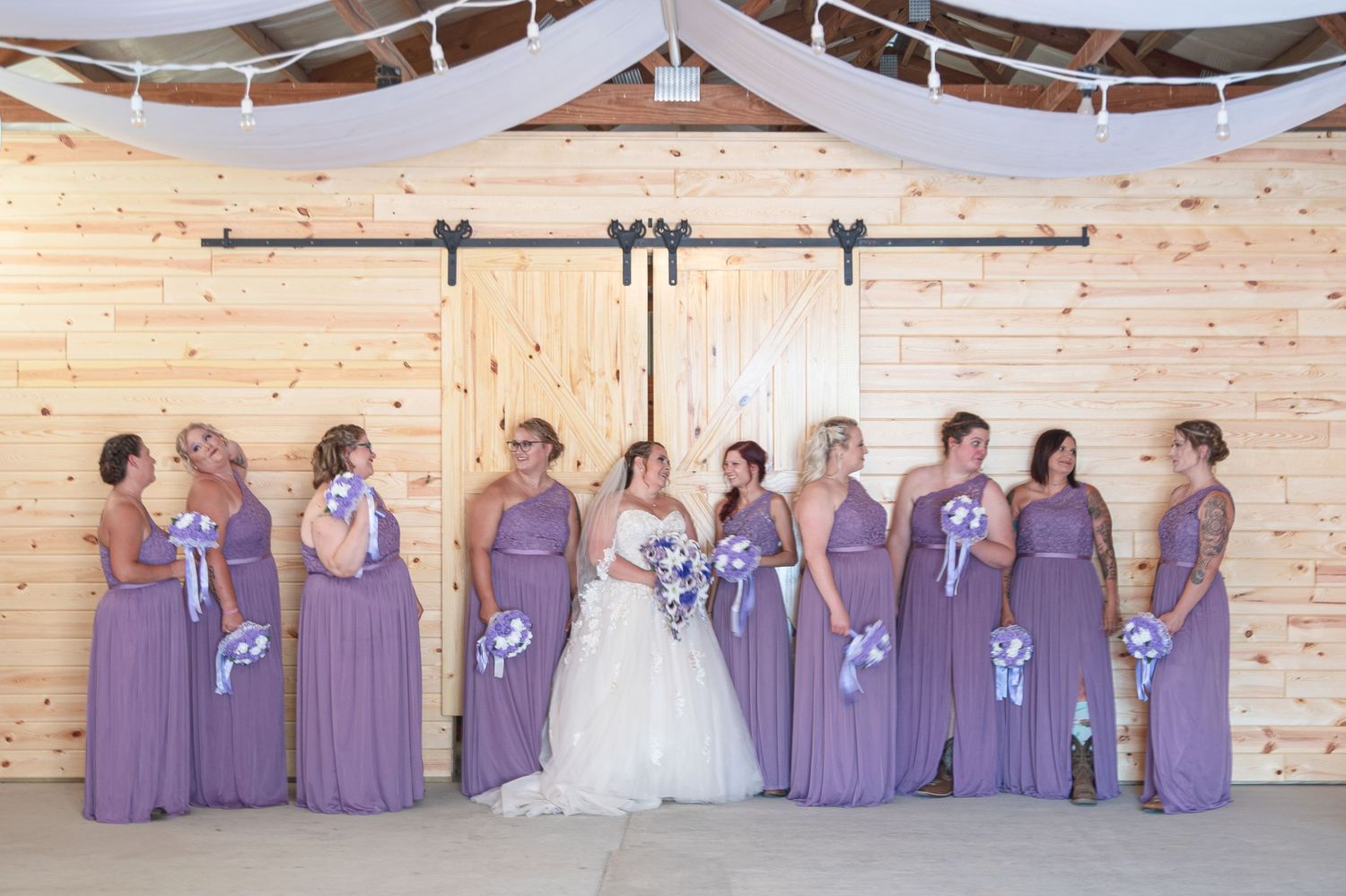 Bride in white gown poses with bridesmaids in lavender dresses inside a rustic wooden barn wedding venue.