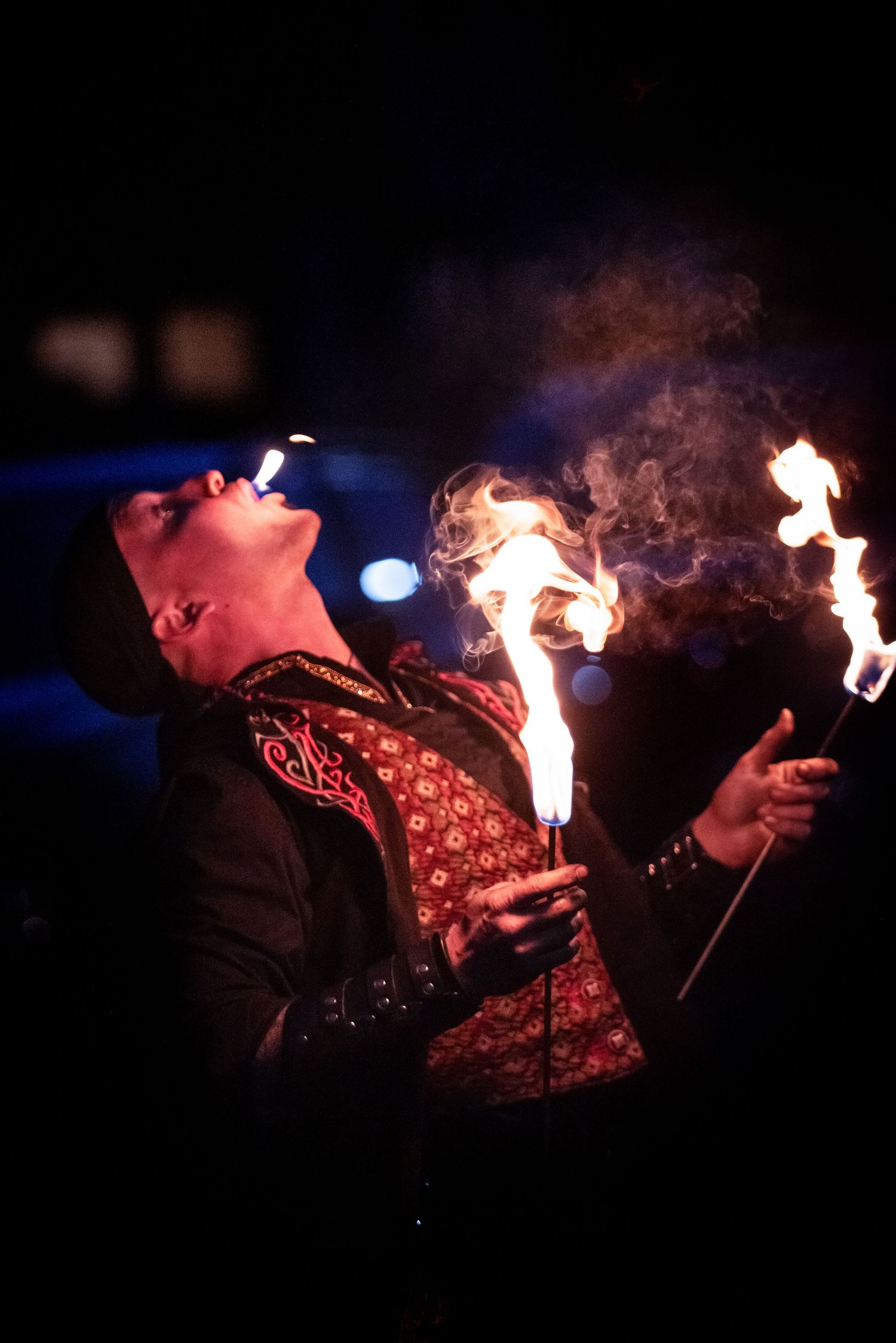 A costumed fire performer exhales smoke while holding flaming torches during a nighttime show.Outside Monster 
Bash Harmony MN.