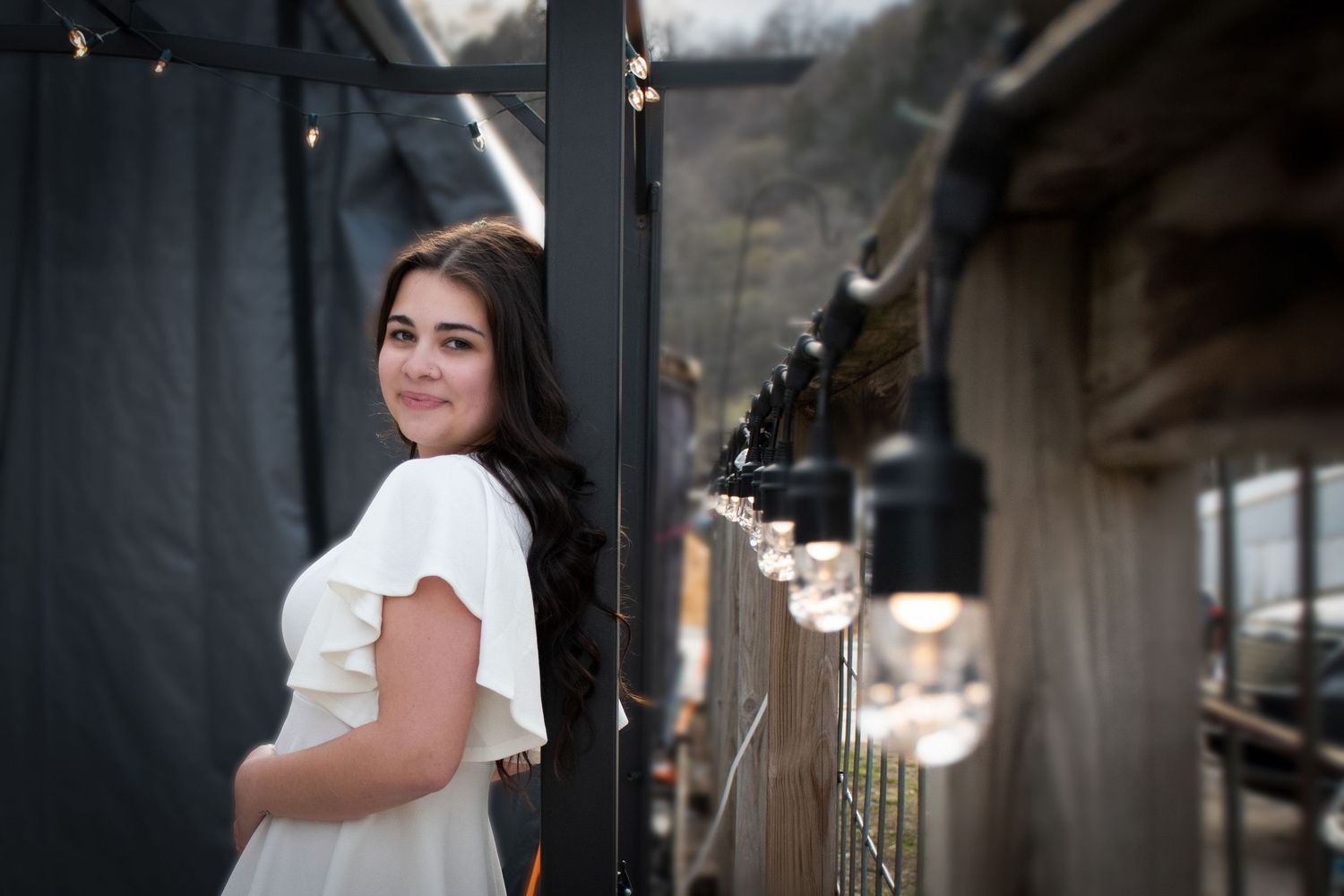 Smiling brunette woman in white ruffled top posing beside string lights outdoors.