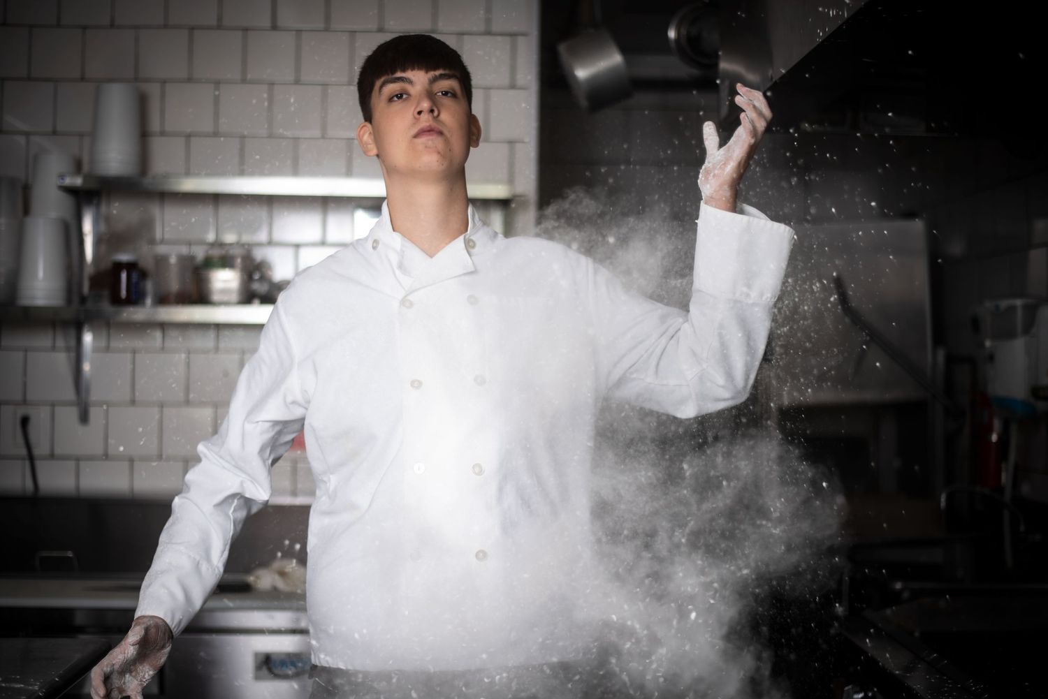 Professional chef in white uniform dramatically tossing flour in a restaurant kitchen.