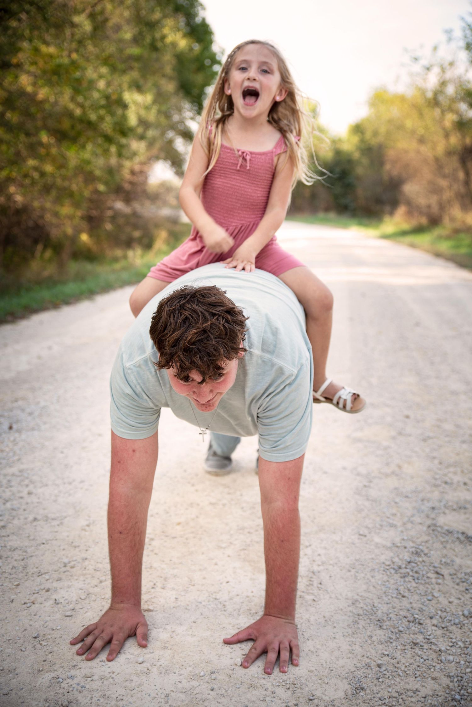 A young girl screams with excitement while sitting on a teenage boy doing push-ups.