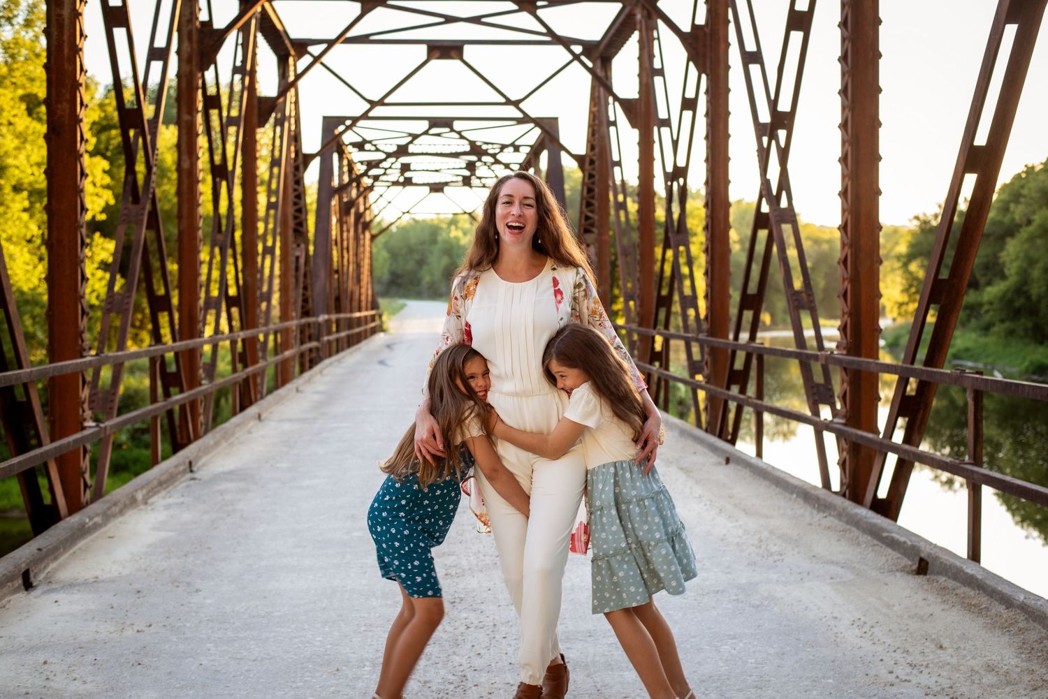 A laughing mother and two daughters playing together on a rustic iron bridge at sunset.