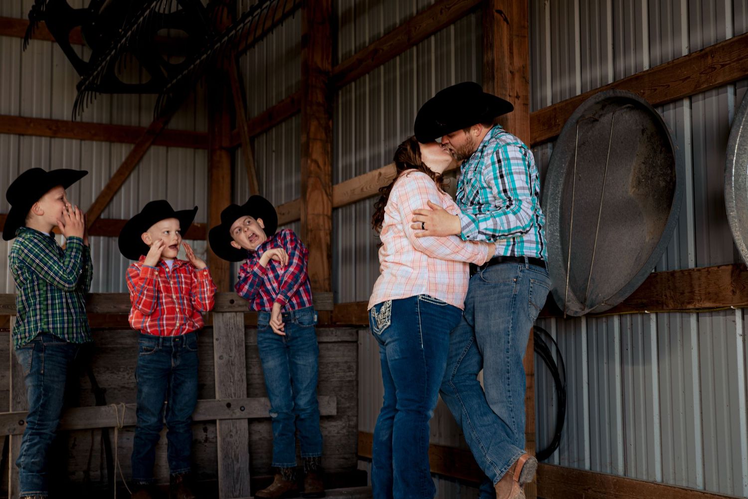Couple sharing a kiss in a rustic barn while three children in cowboy hats look away playfully.