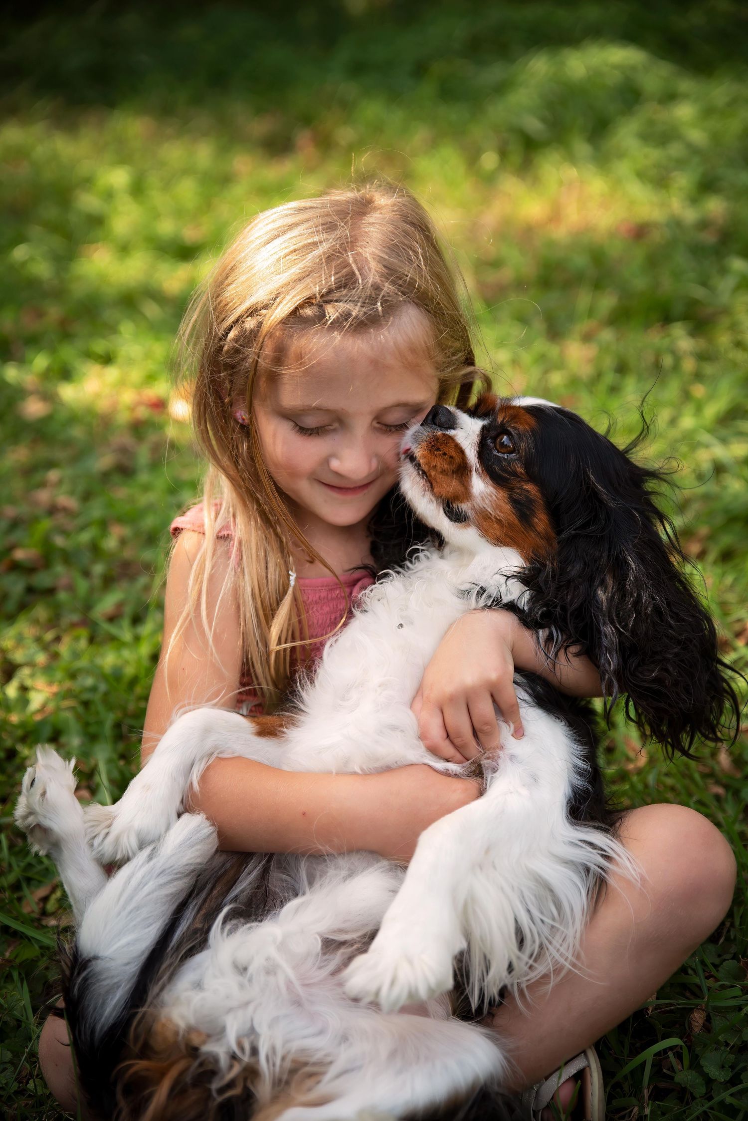 A smiling girl sits on grass as a Cavalier King Charles Spaniel gives her a kiss.