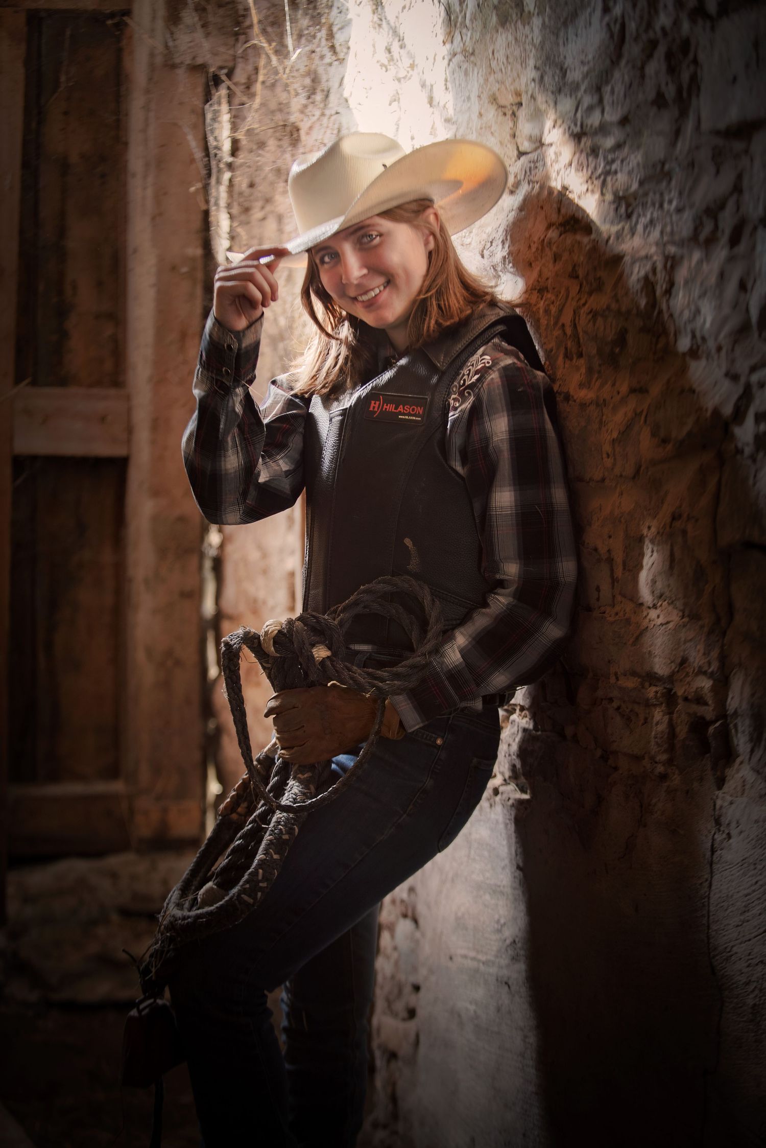 Confident cowgirl in white hat and plaid shirt leaning against a rustic stone wall.