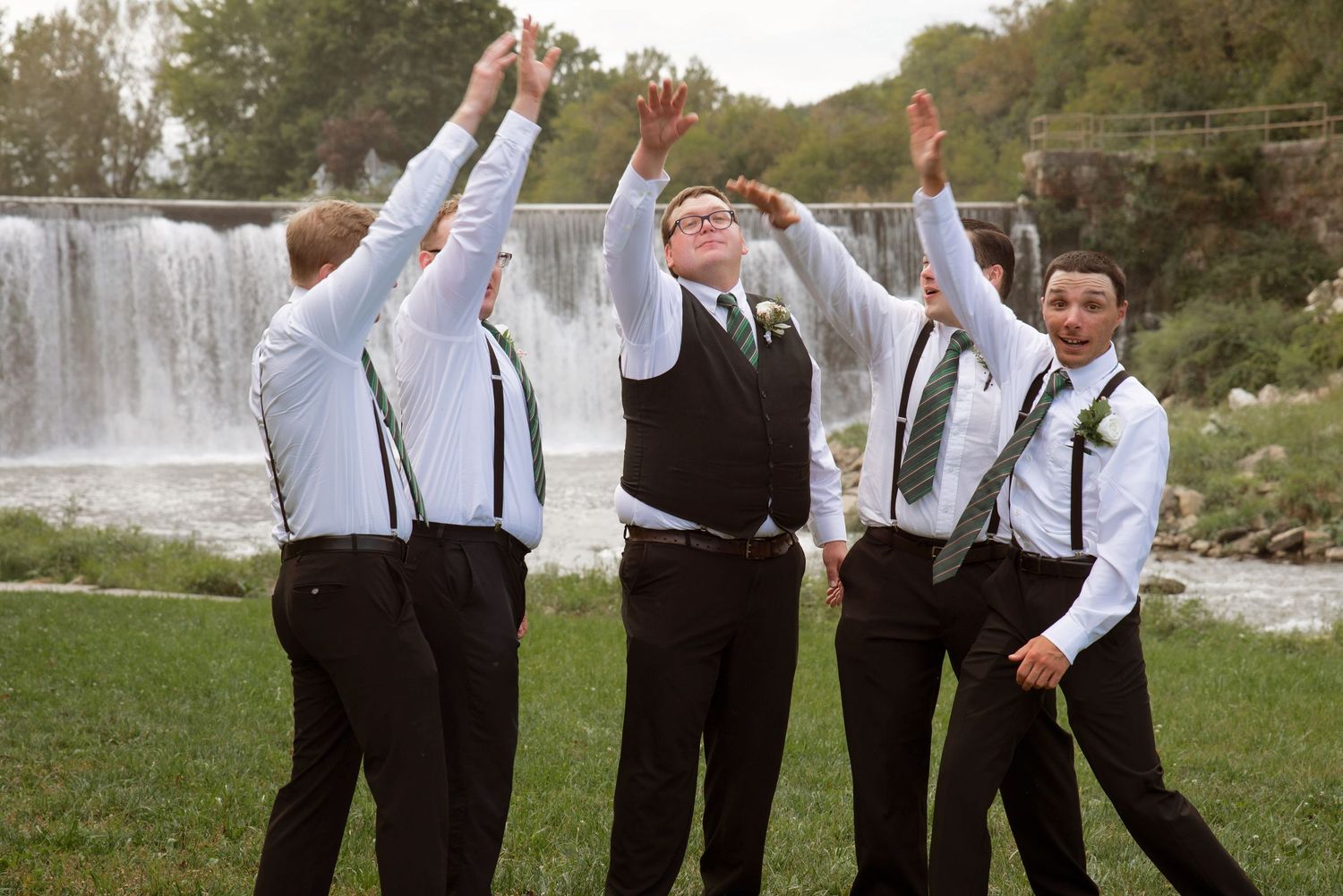 Groom and groomsmen in black suspenders raise arms joyfully in front of scenic waterfall backdrop in Lanesboro MN.