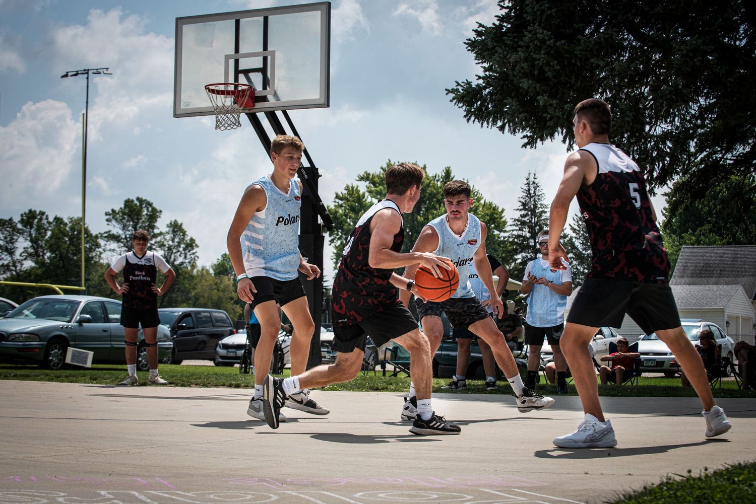 Group of young men playing competitive outdoor Rucker League basketball near a regulation backboard on a sunny day.