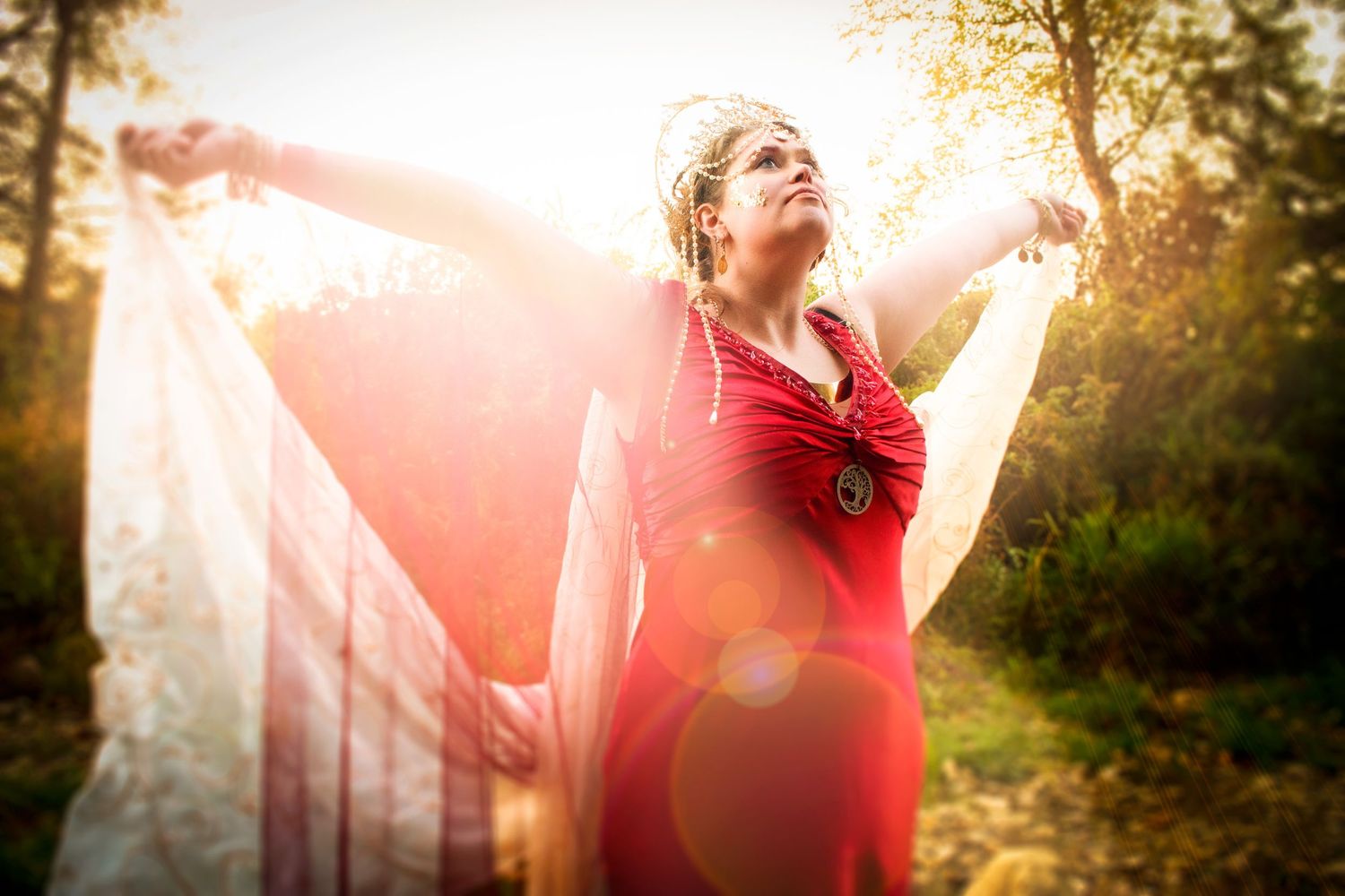 Woman in red dress with flowing white fabric outstretched arms in golden sunlit forest.