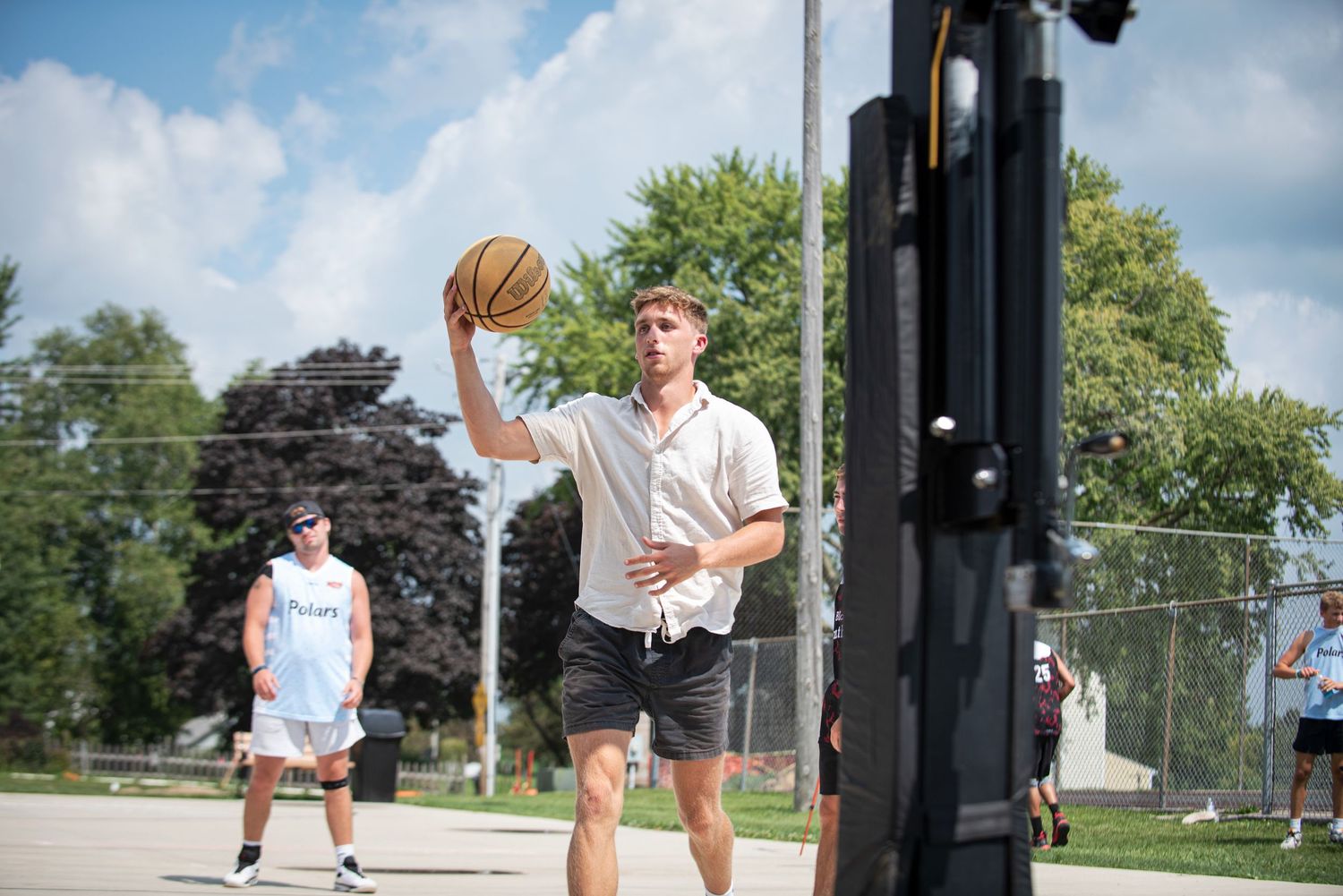 Young man in casual clothes shooting a basketball on an outdoor court with trees in the background.