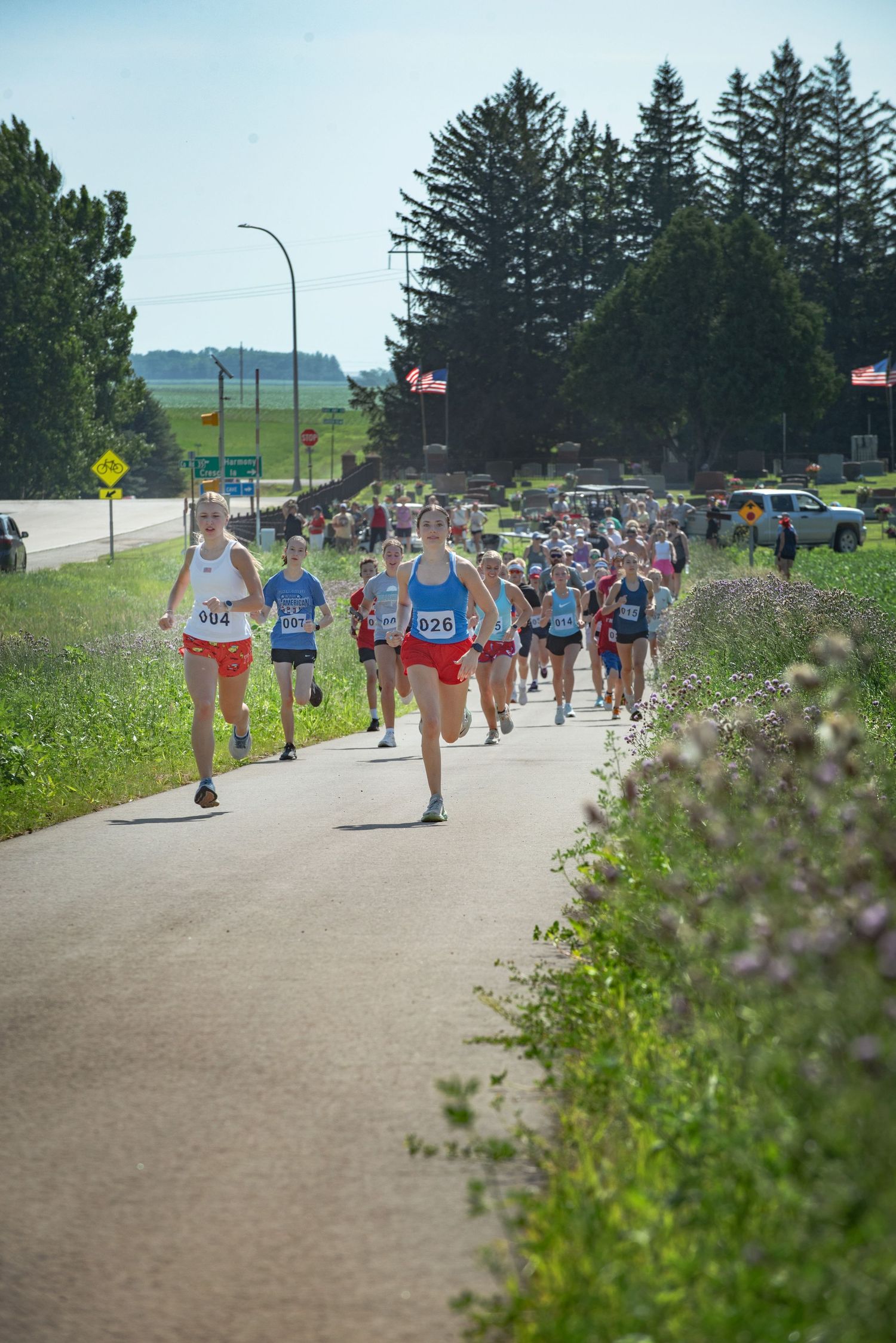 Large group of runners competing in an outdoor road race along a paved path on a bright sunny day at Harmony MN 4th of July Celebration.