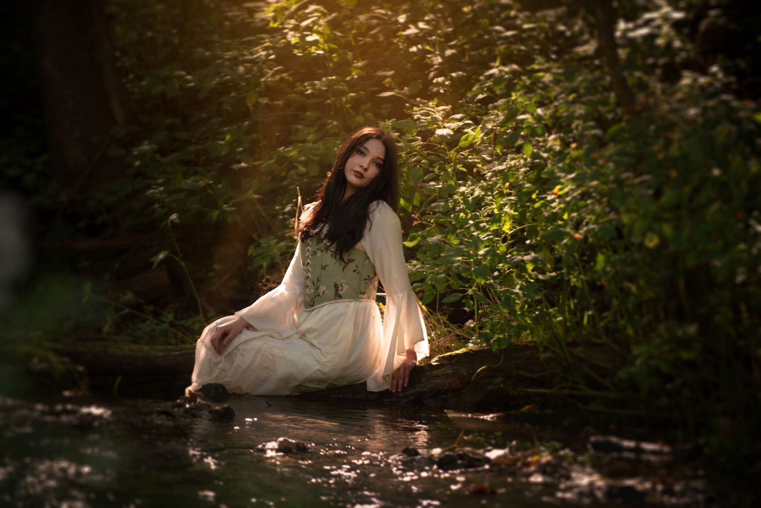 Beautiful woman in white dress sitting peacefully by a forest stream in golden sunlight.