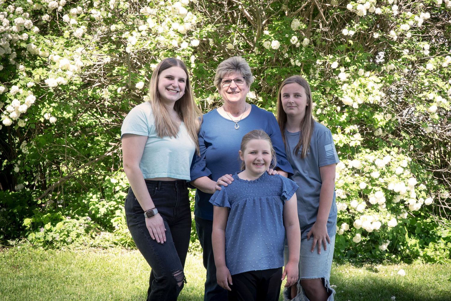 Three generations of women smiling together in front of blooming white flower bushes.