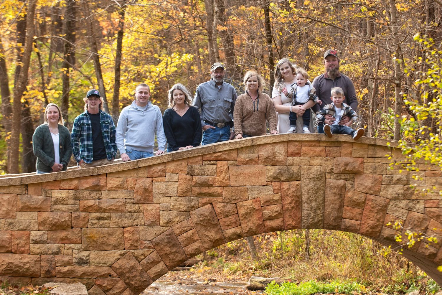 Extended family group portrait on a stone arch bridge surrounded by vibrant autumn foliage in a wooded park.