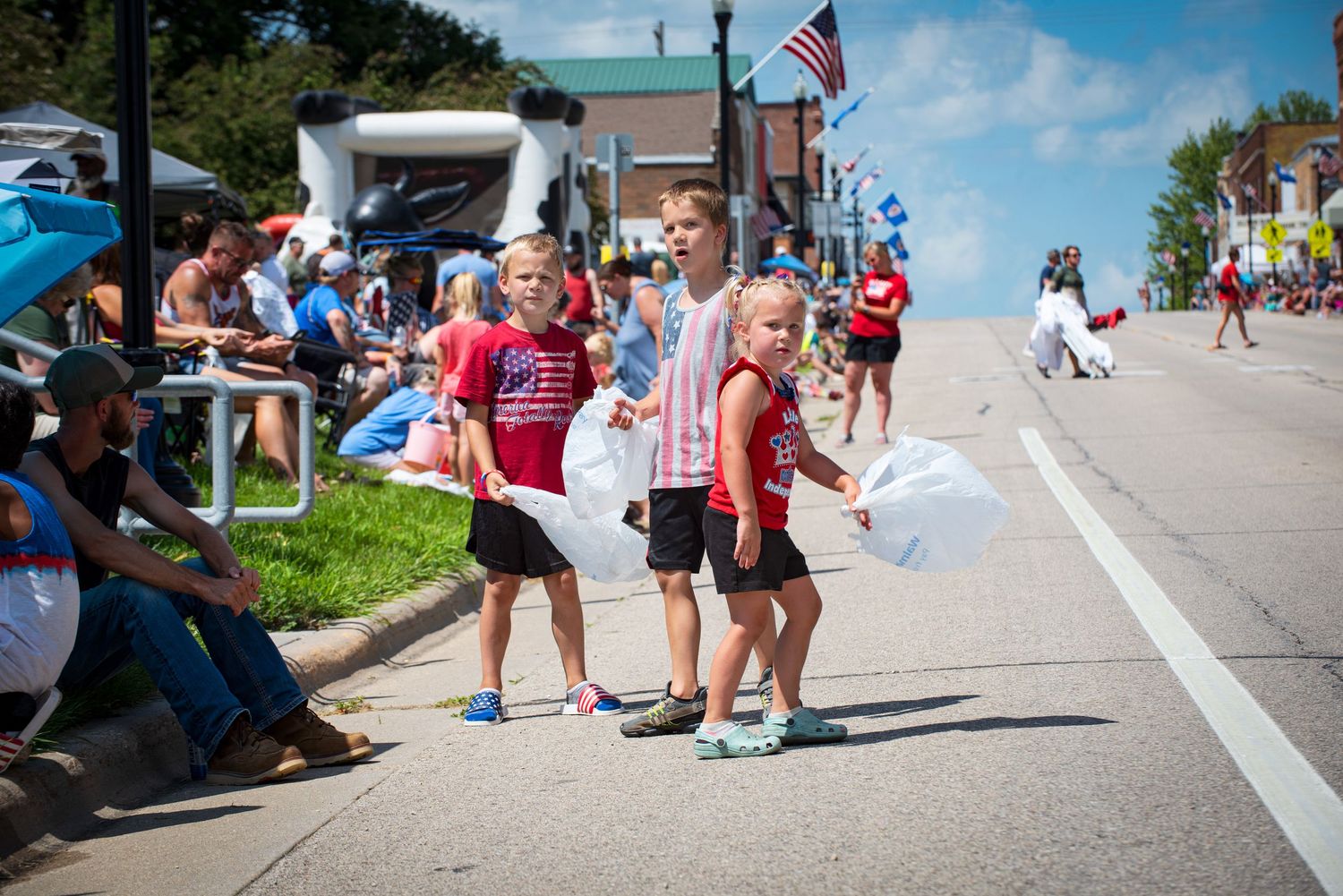 Children in red shirts hand out bags to spectators during a festive Fourth of July parade on a sunny small-town street. At Harmony MN.