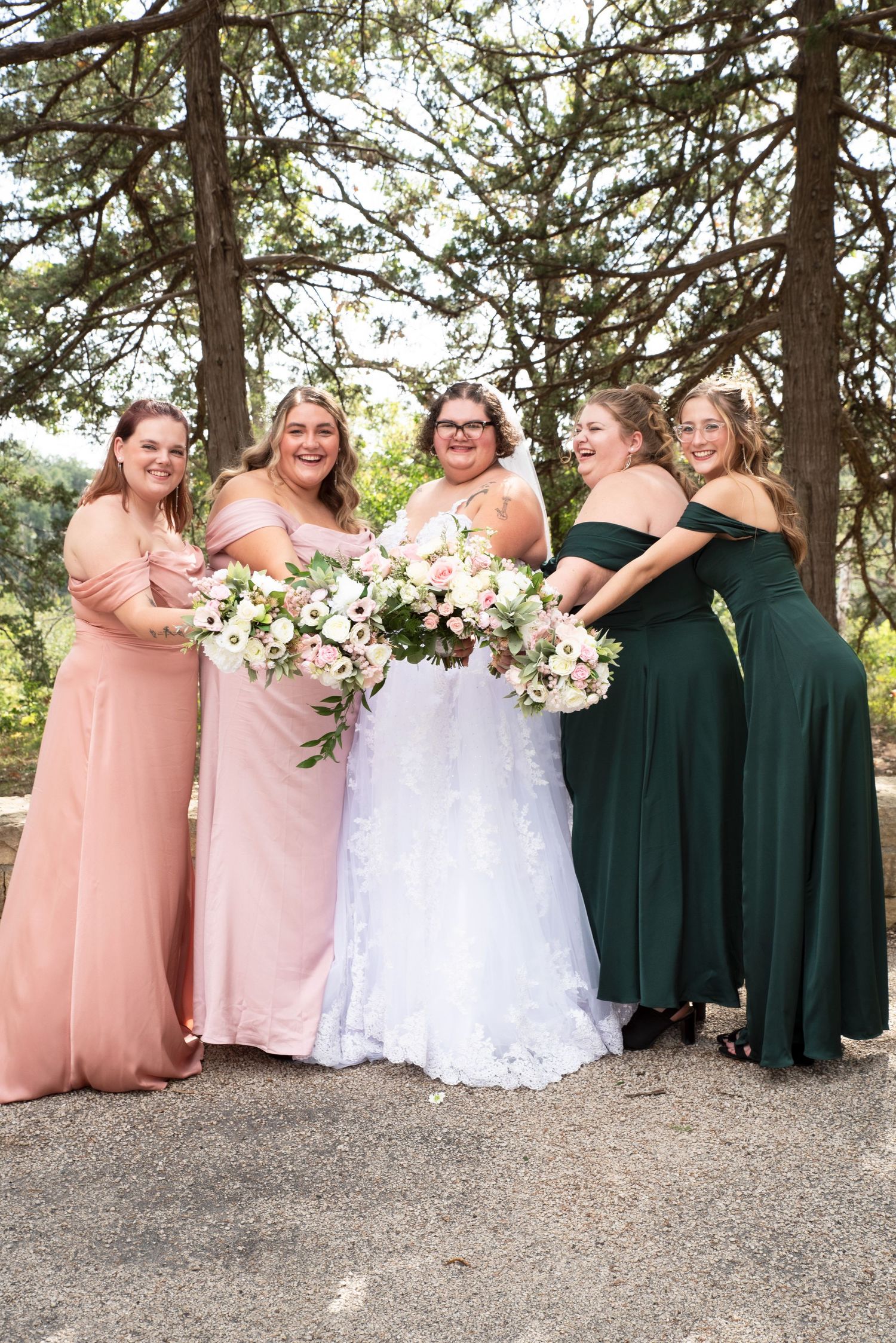 Bride with four bridesmaids in pink and green off-shoulder gowns holding bouquets under pine trees in Lanesboro MN.