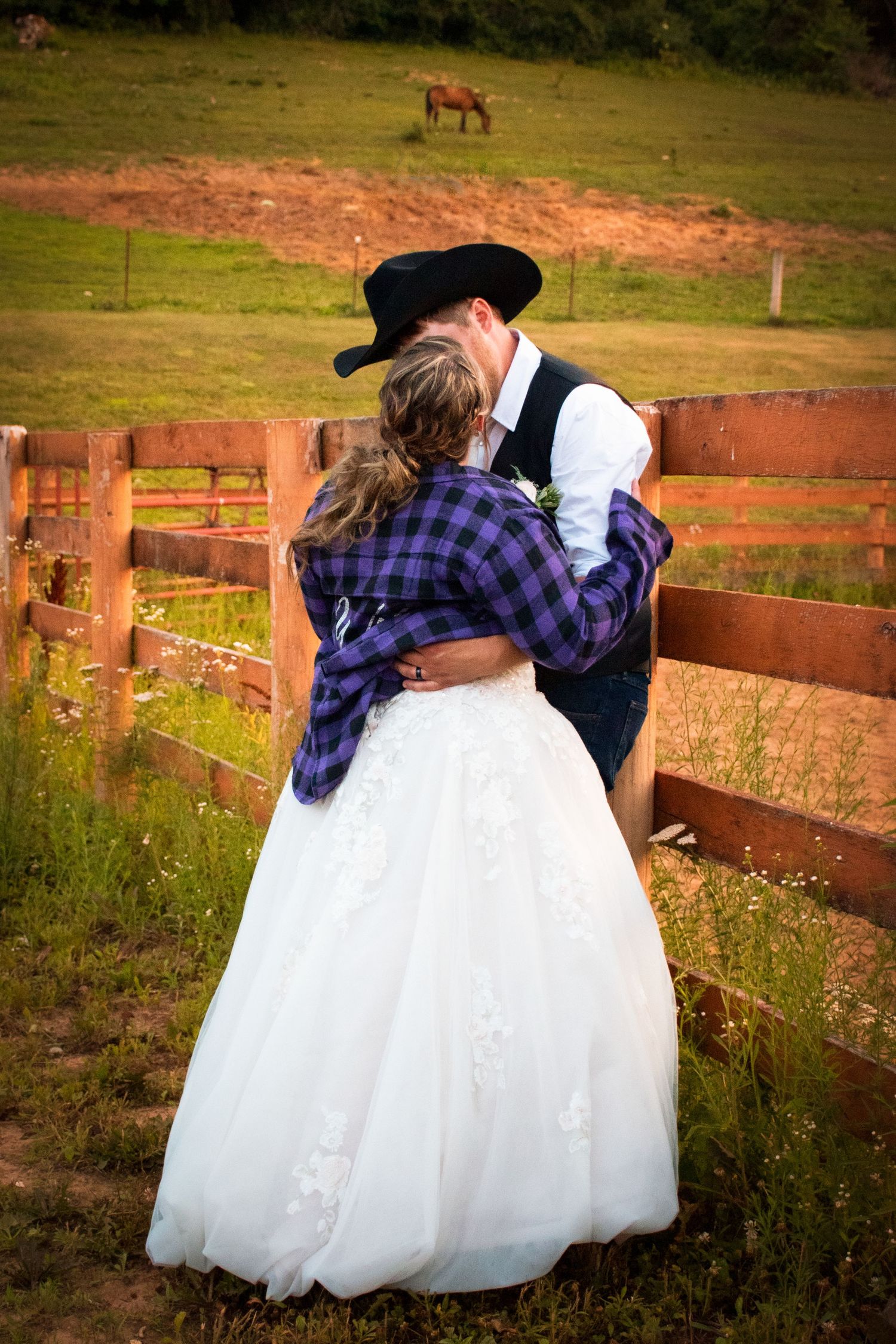 Bride and groom share romantic embrace by wooden fence with horse grazing in green pasture behind.