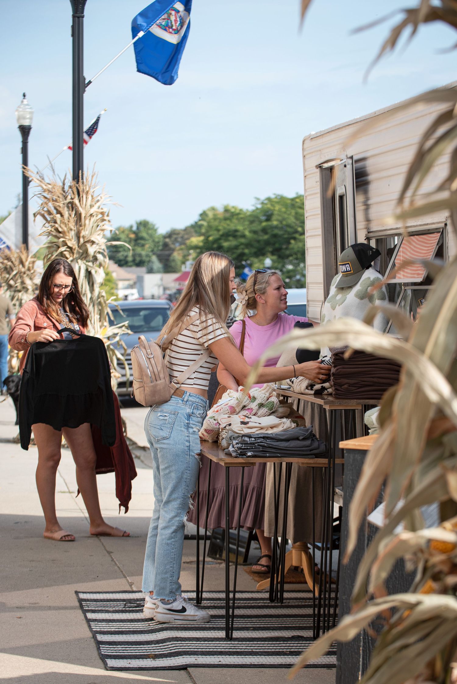 Women shopping at an outdoor clothing vendor on a sunny small-town sidewalk in Harmony MN.