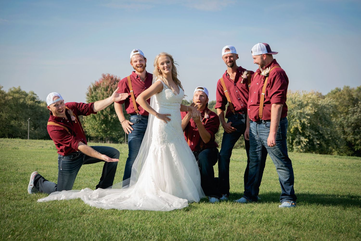Bride in white mermaid gown poses with groomsmen in maroon shirts and baseball caps on sunny outdoor lawn.