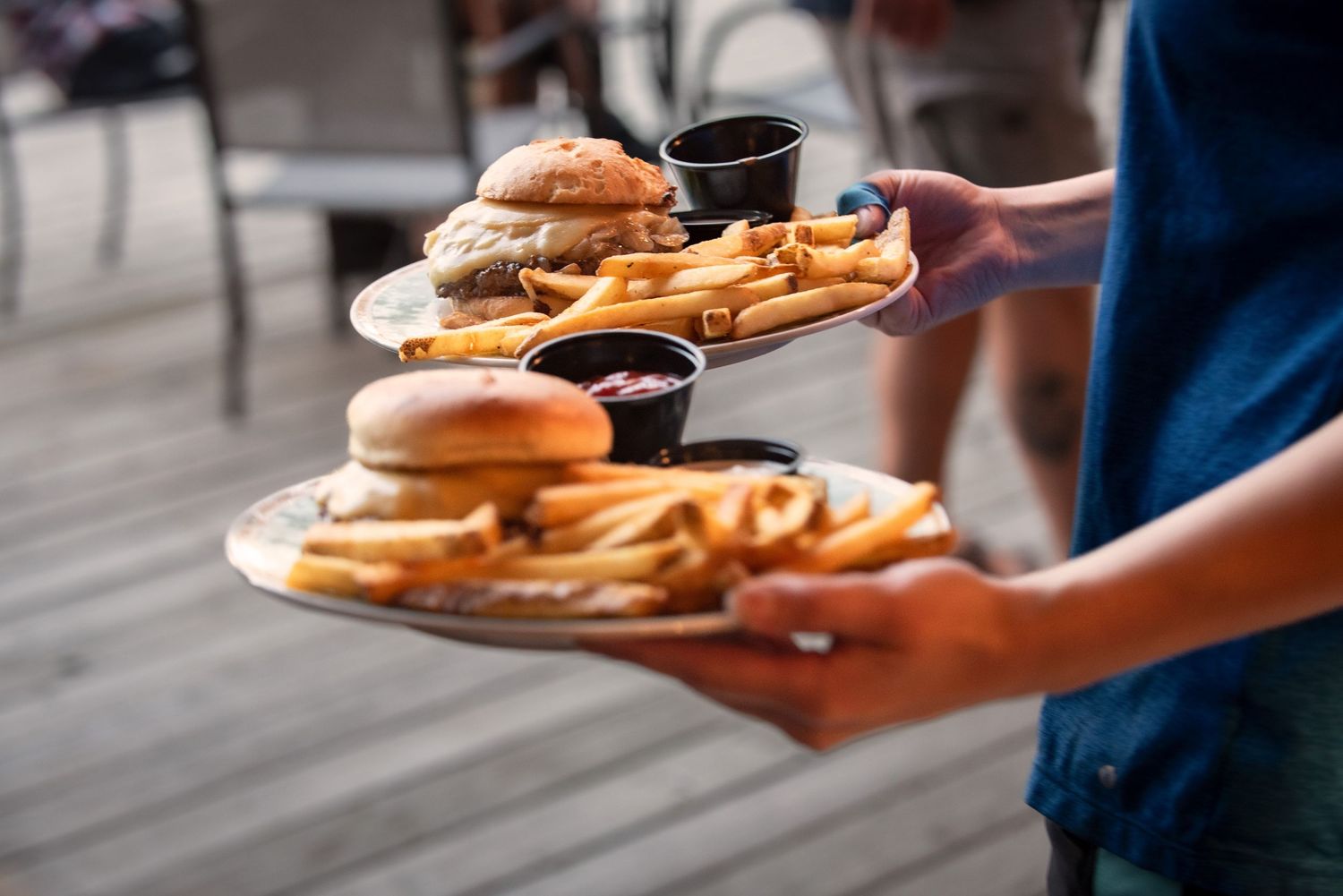 Server carrying two plates of burgers and french fries with dipping sauces on outdoor deck at Estelle's Eatery.