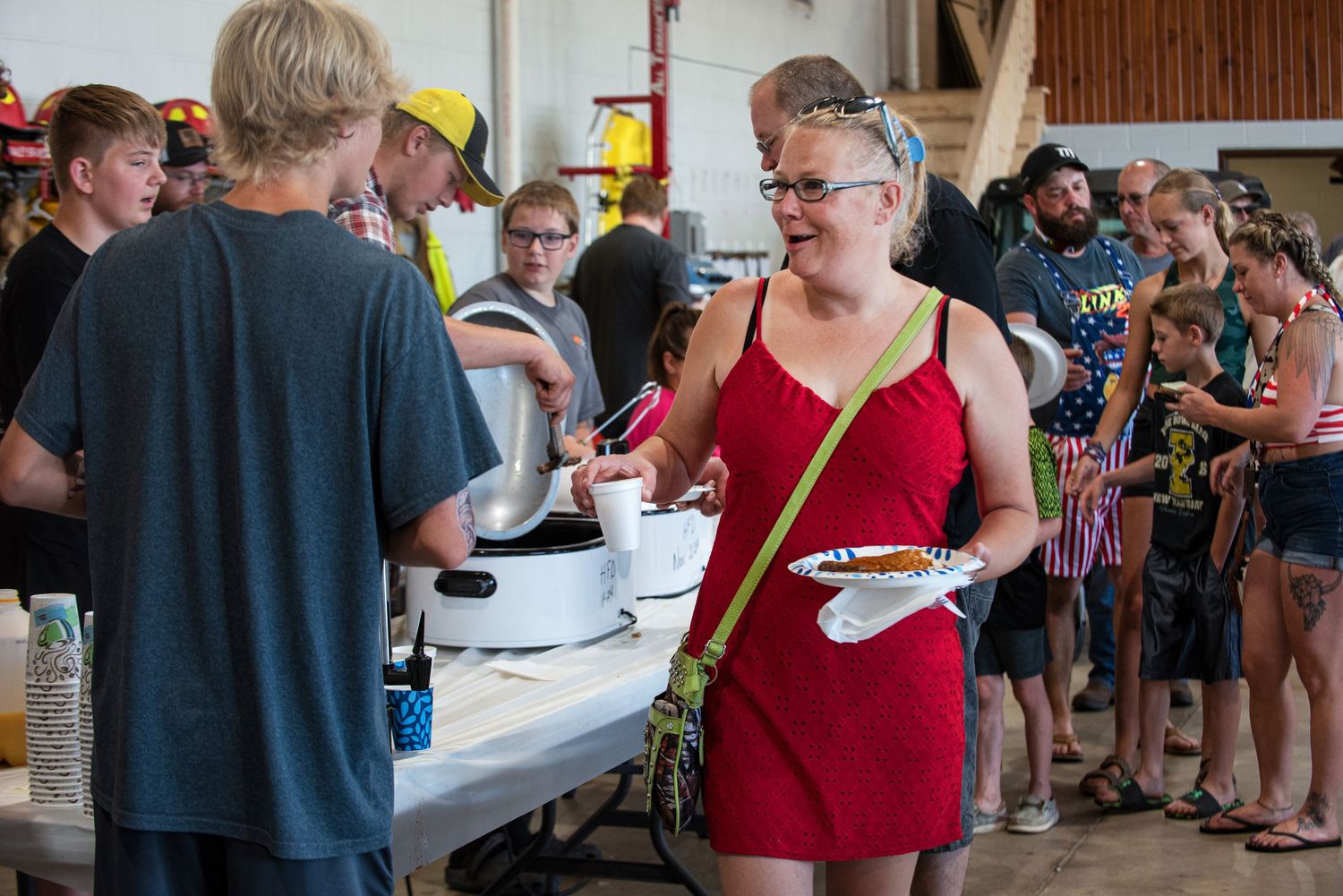 A woman in a red dress serves food to guests in a long buffet line at a community gathering. 4th of July Celebration in Harmony MN.