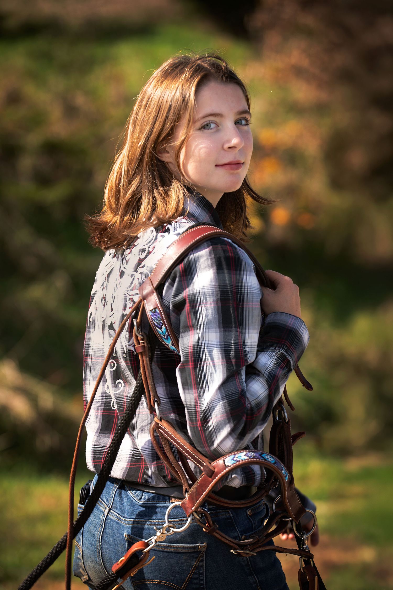 Young woman in plaid shirt holding horse bridle, looking back over shoulder in autumn setting.