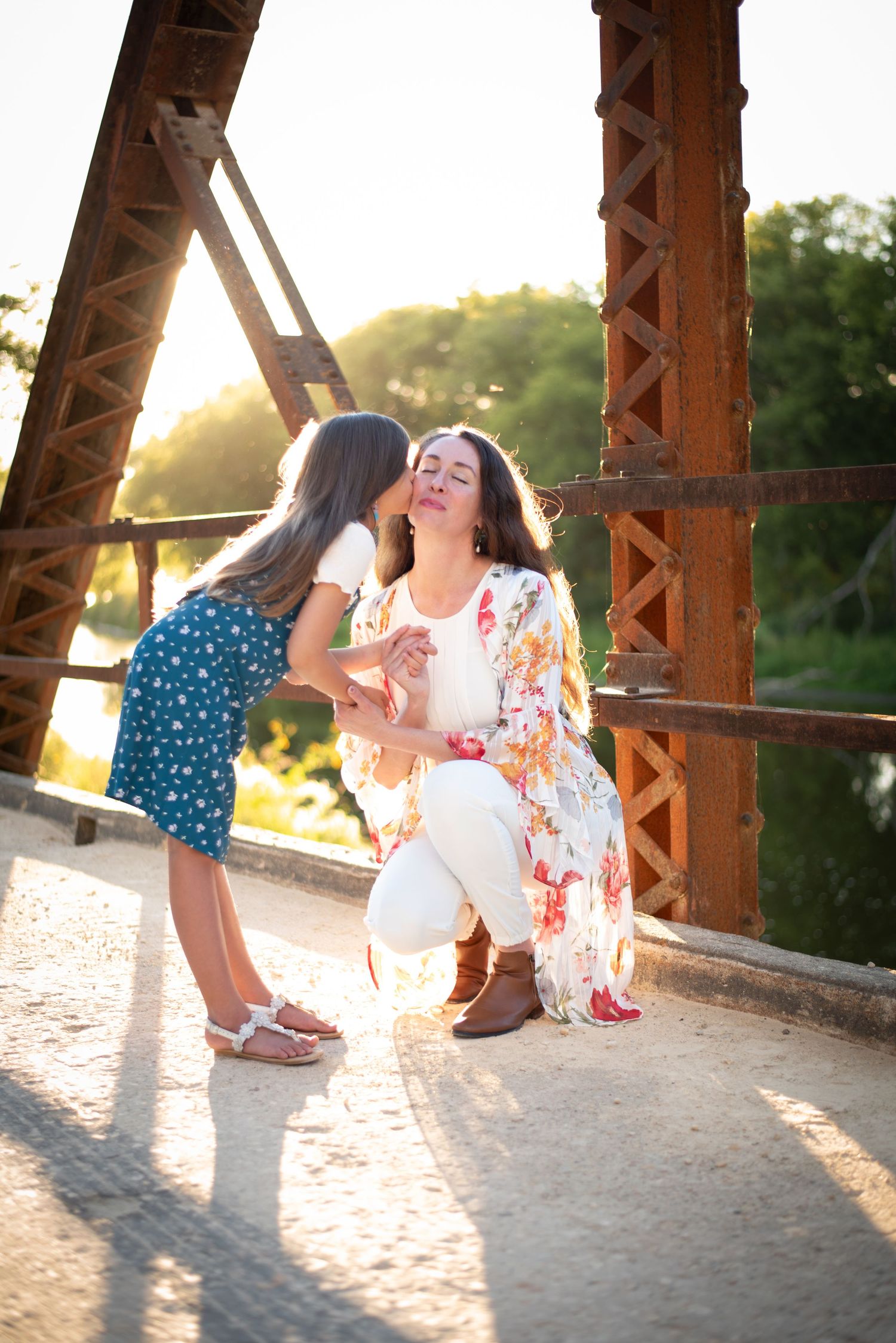 A young girl kissing her mothers cheek on a sunlit rustic bridge.
