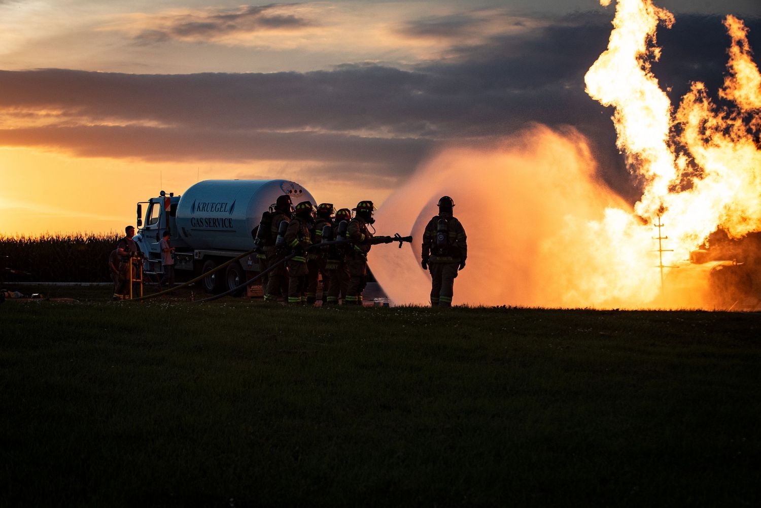 Firefighters battling a massive practice fire at sunset with silhouettes against flames.