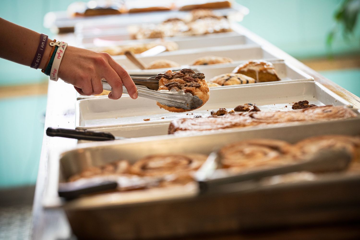 Hand selecting baked pastries and cinnamon rolls from a buffet serving tray at Gabby Lou's