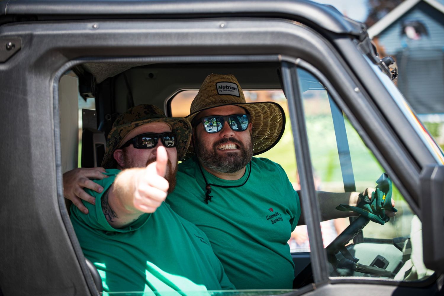Two smiling men in green shirts and hats give a thumbs up from inside a vehicle during a parade. at Harmony MN 4th of July Celebration.