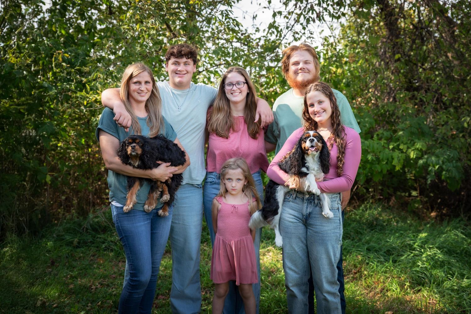 A cheerful family of five poses outdoors with two adorable dogs in lush greenery.