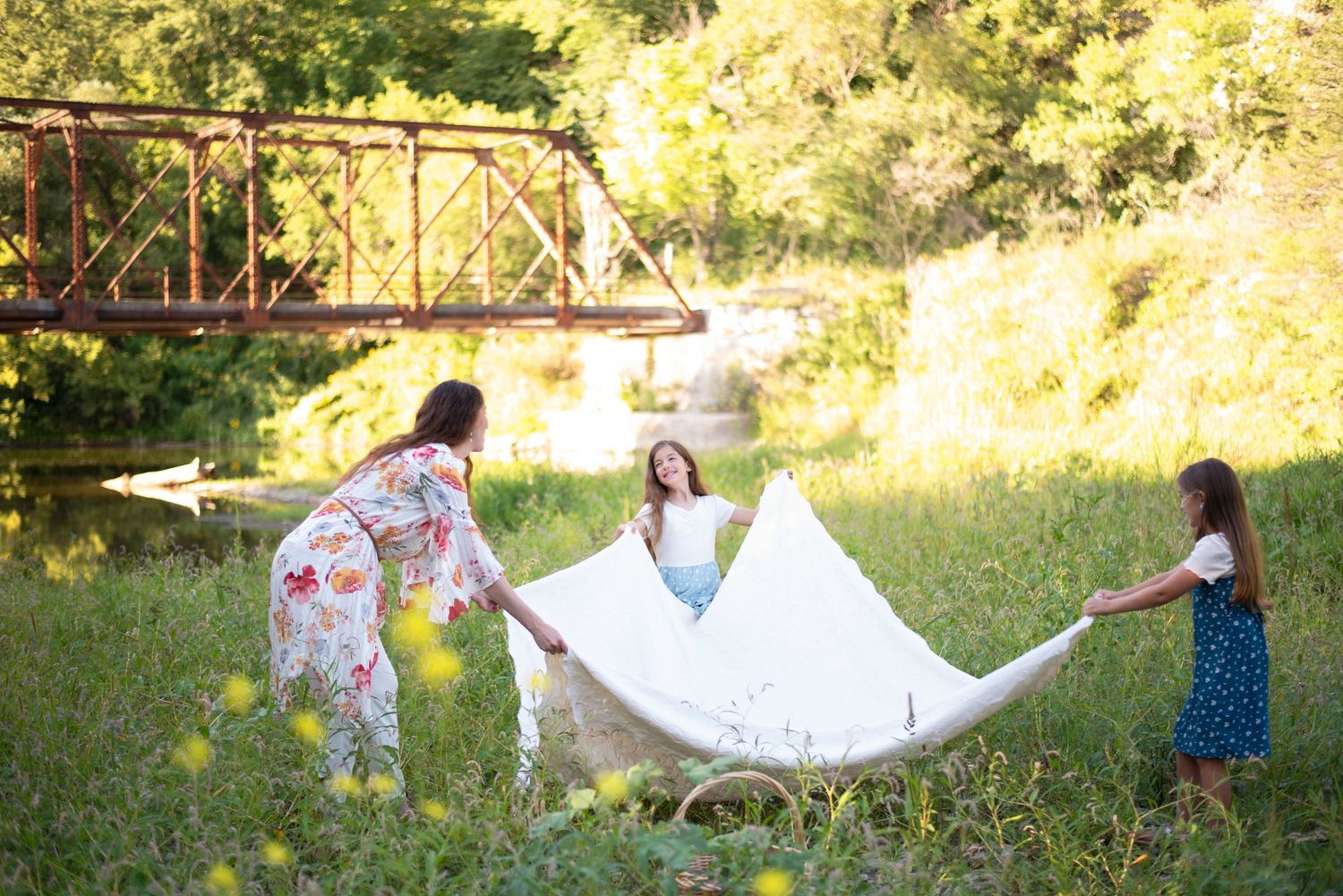 A mother and two daughters playfully shaking out a white blanket in a sunny meadow.