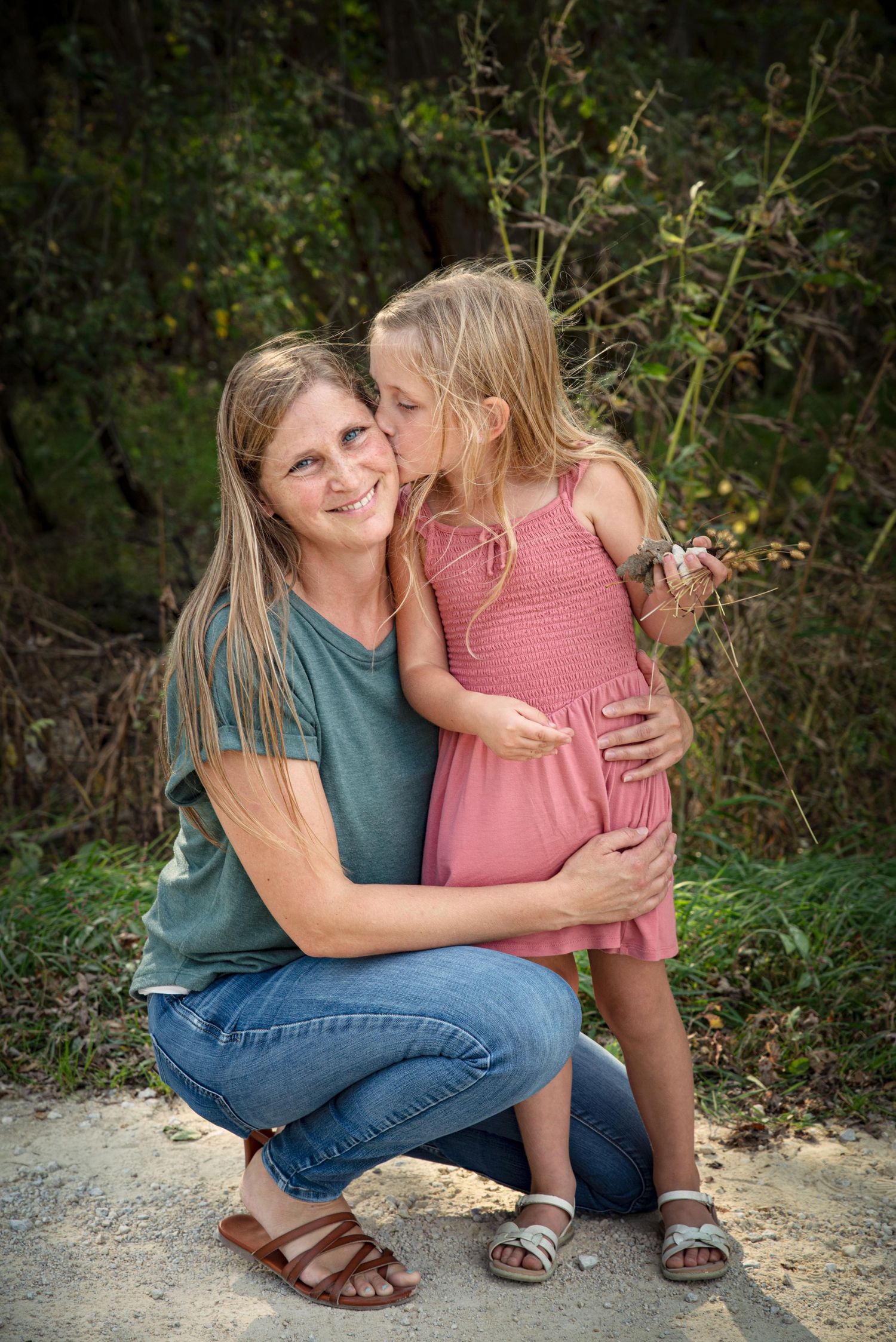 A smiling woman crouches as a young blonde girl in a pink dress gives her a sweet kiss on the cheek.