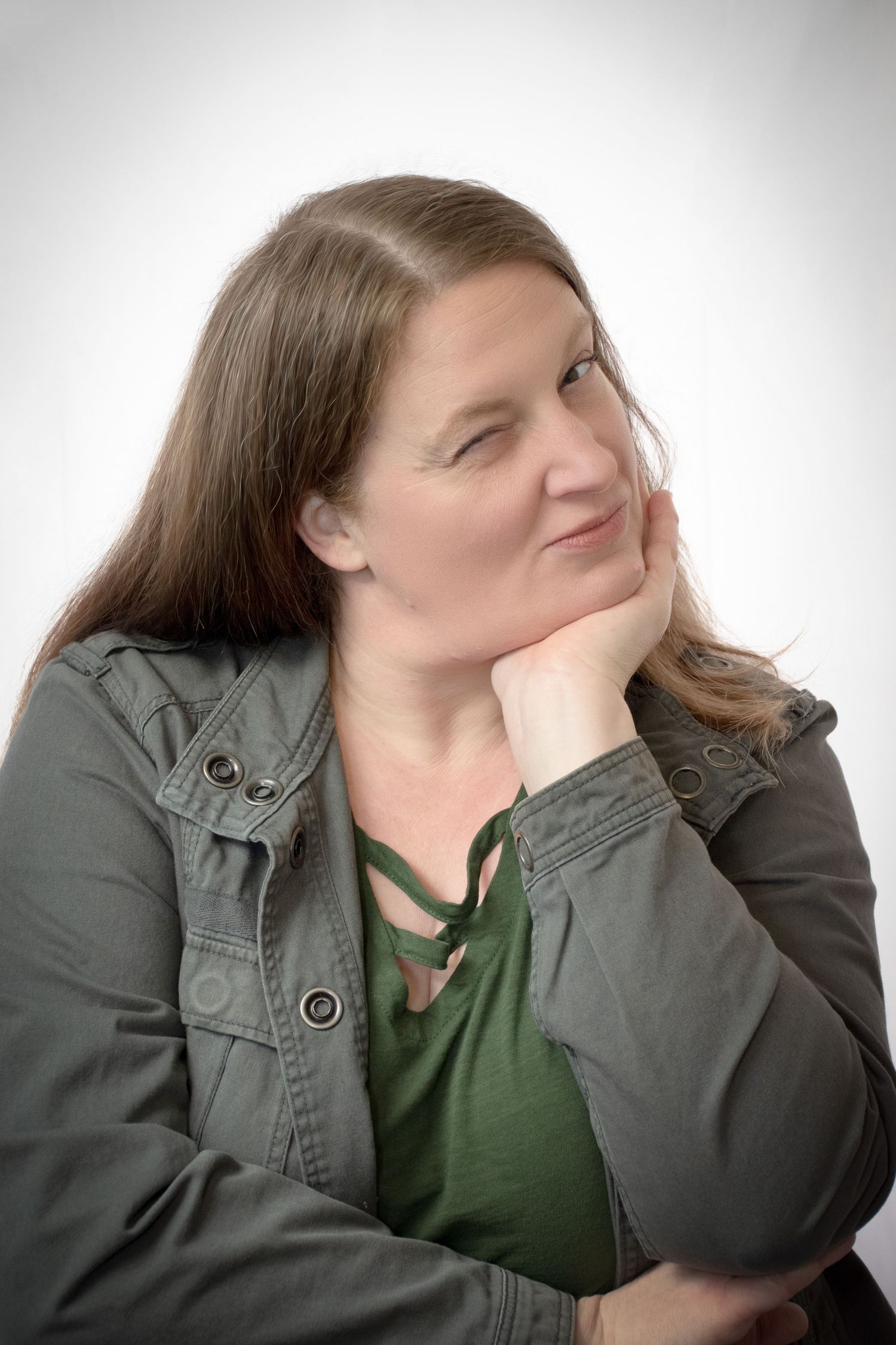 Woman with long brown hair rests chin on hand, wearing an olive green jacket, looking thoughtfully to the side.