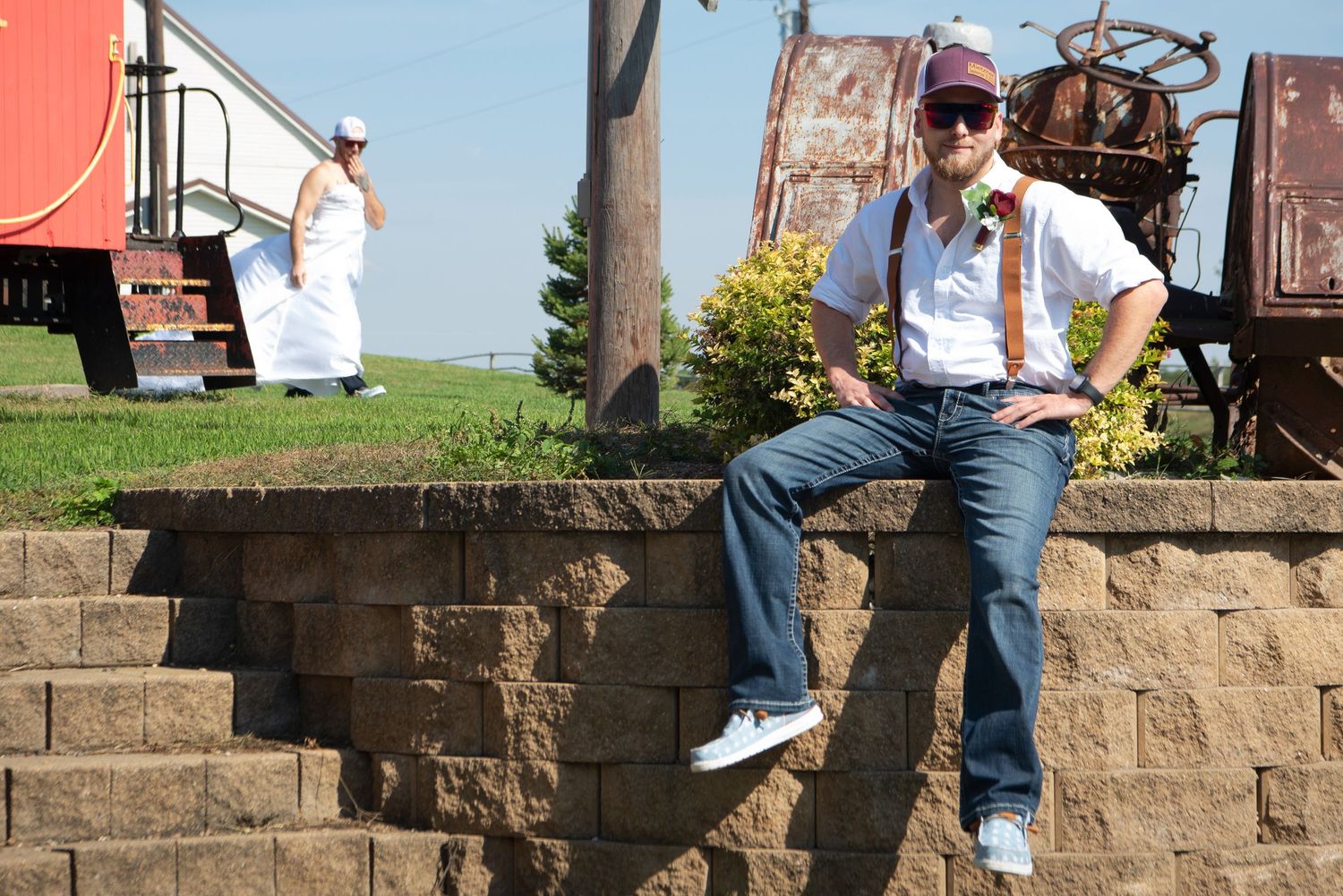 Groom in white shirt, suspenders, and sunglasses sits on stone wall outdoors near vintage train cars on wedding day.