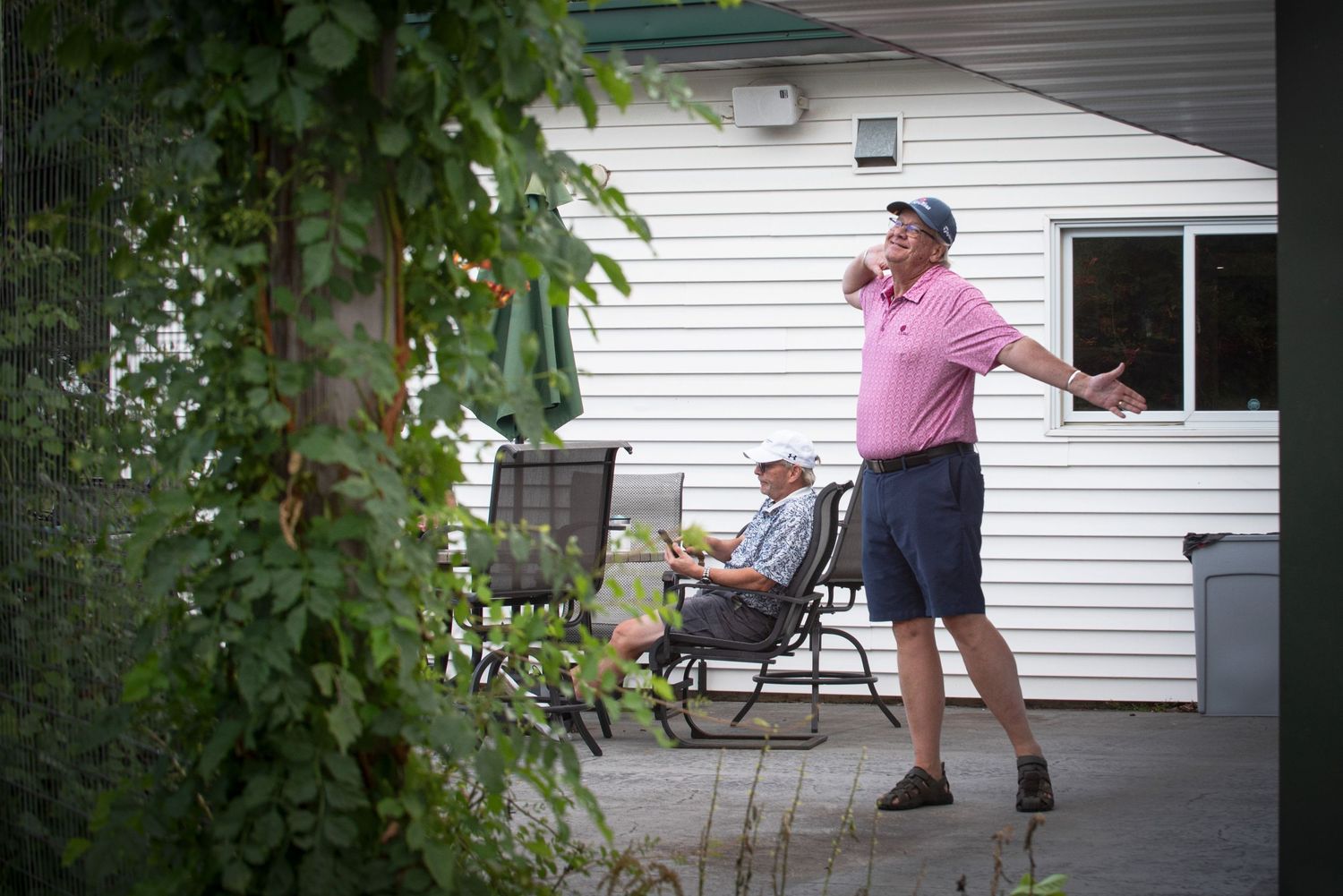 Two men relaxing on a backyard patio surrounded by lush green vines and trees.