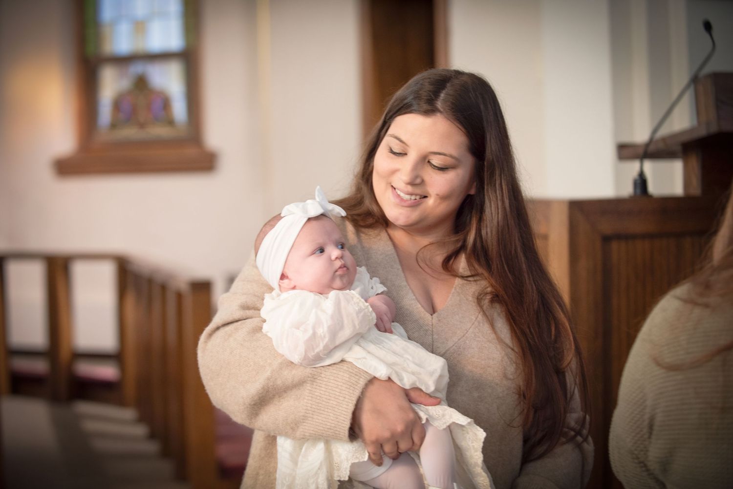 A smiling young woman holds a newborn baby in a white christening gown inside a church.