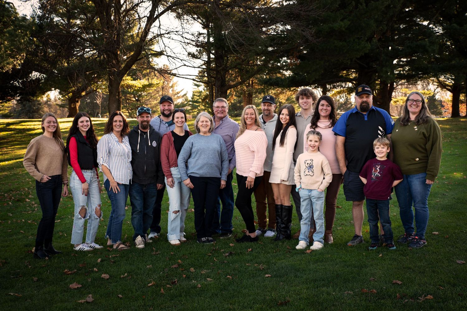 A large multigenerational family poses together on a green lawn with tall autumn trees in the background.