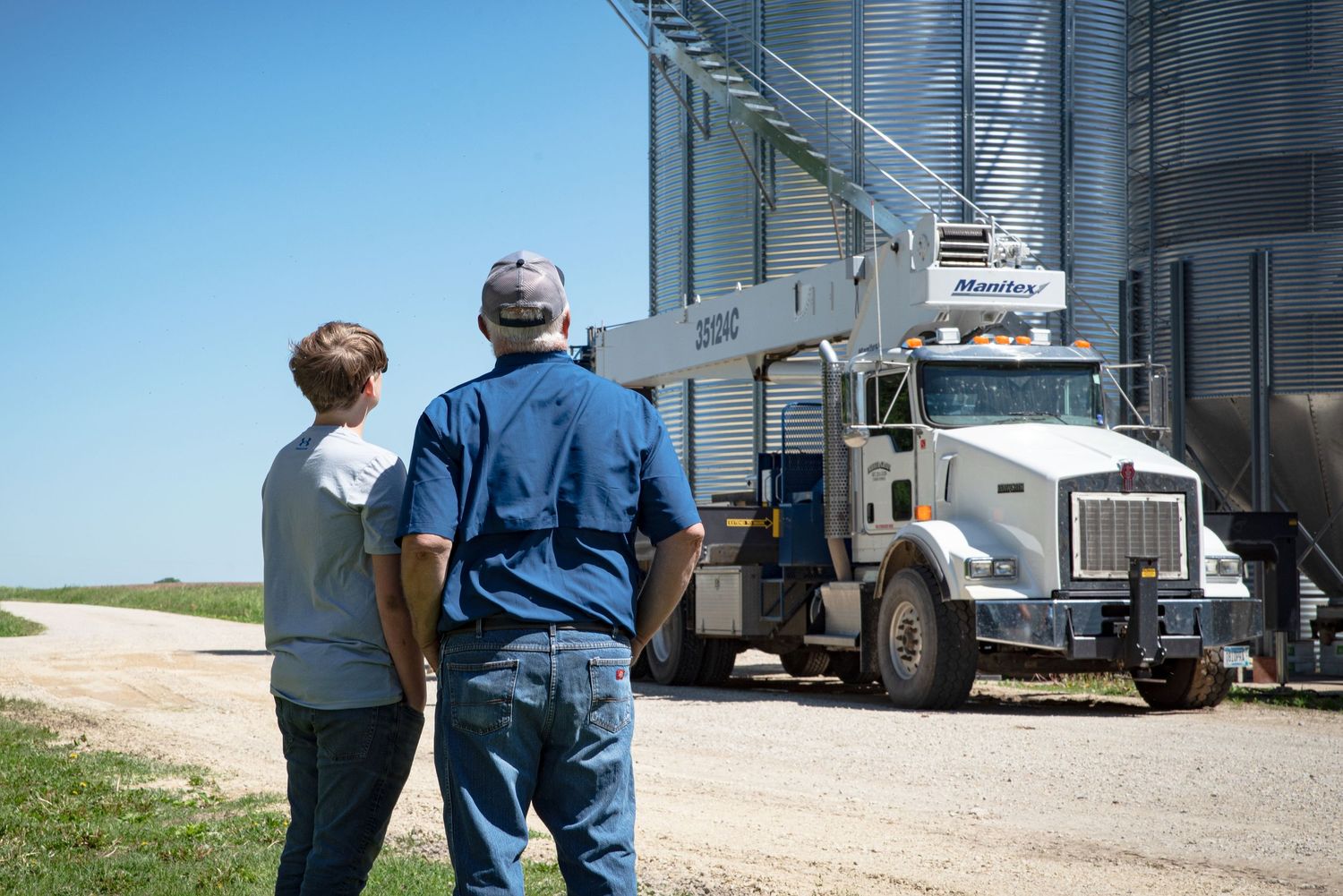 Two people watching a large grain truck unload at a farm storage facility.